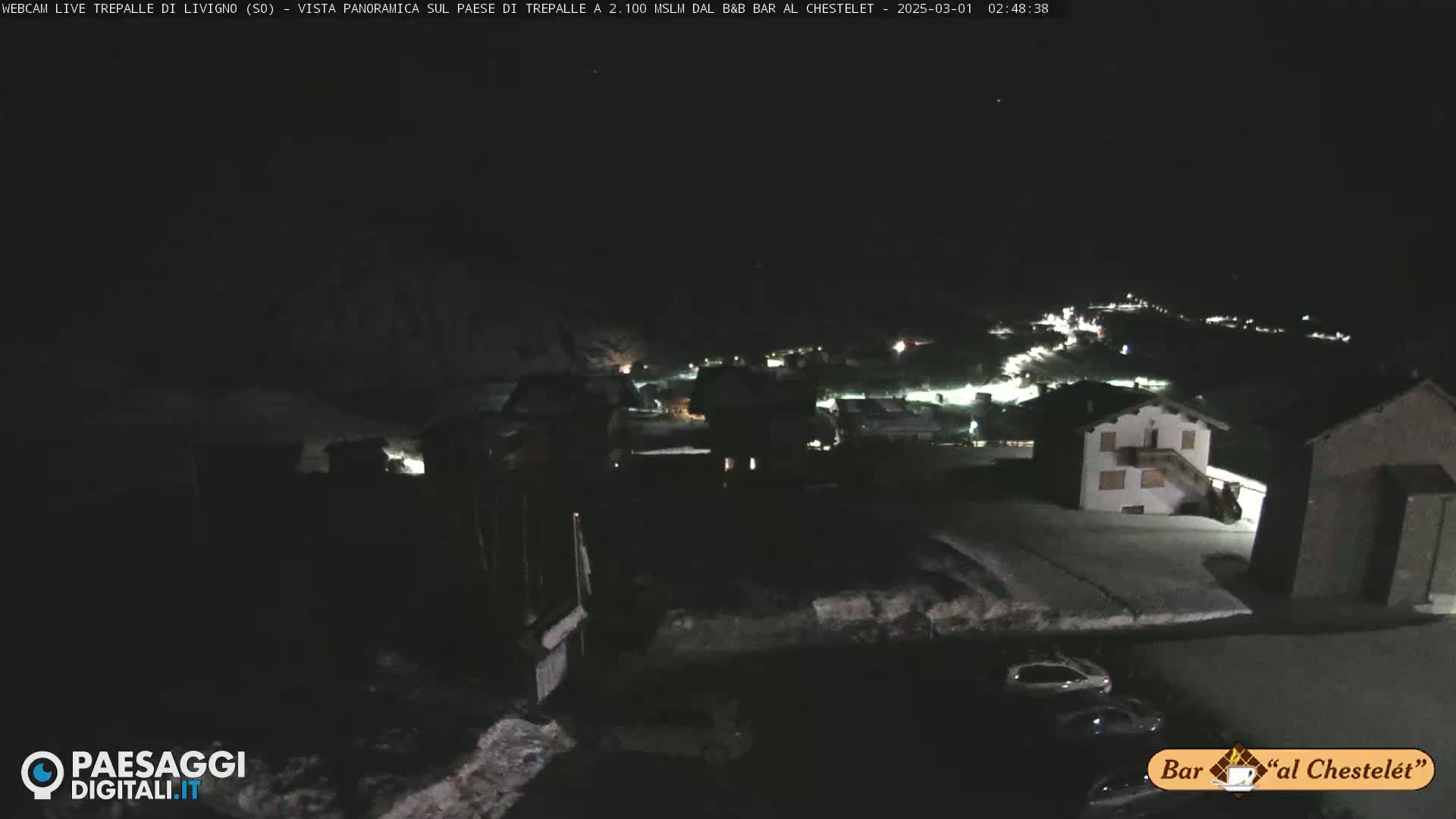 A nighttime, snow-covered village is dimly lit under a dark sky.