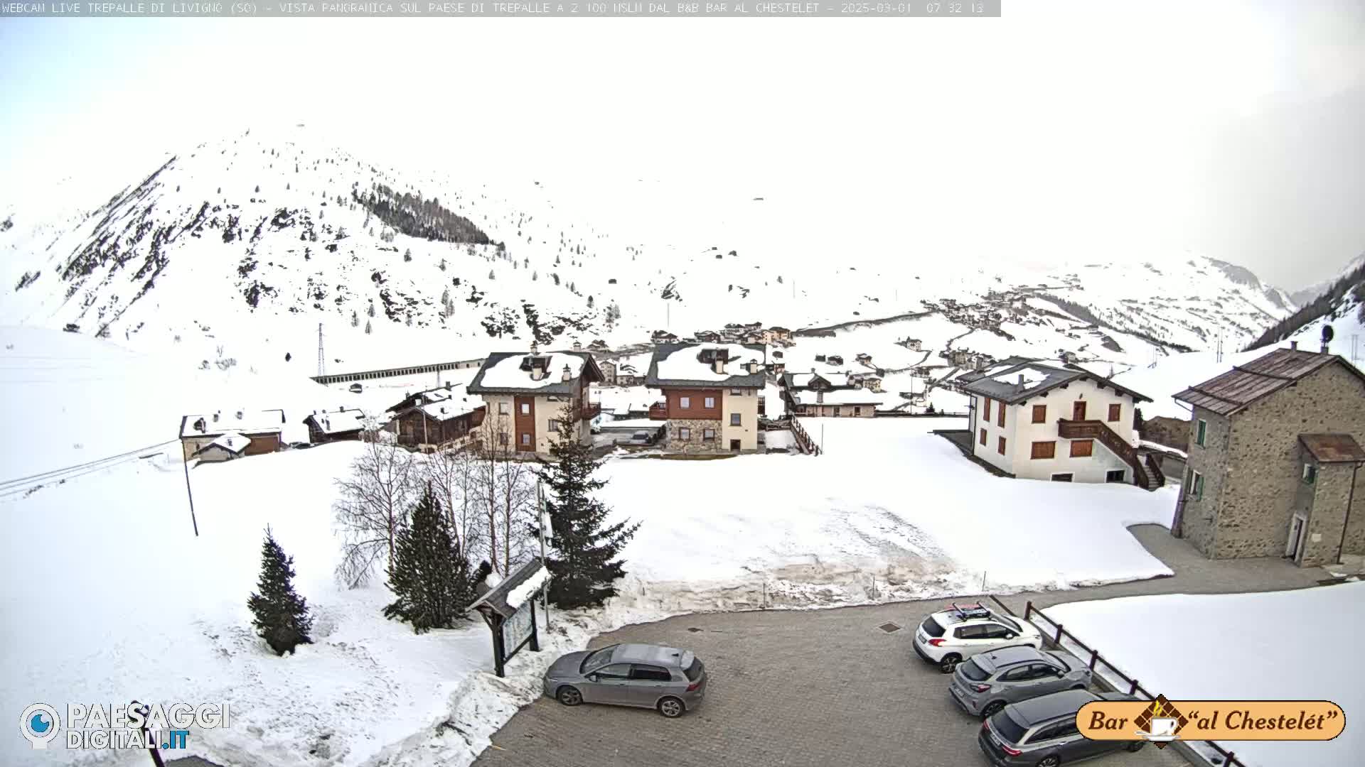 A snow-covered village nestled in a mountain valley under an overcast sky, with several cars parked in a lot.