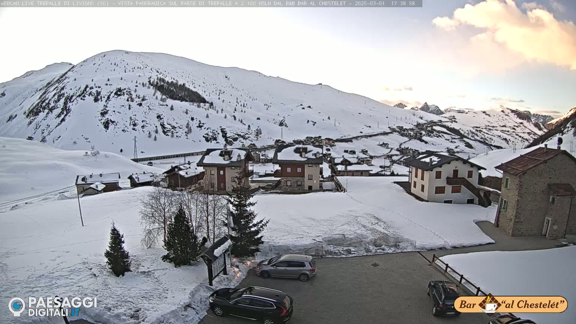 A snow-covered village nestled in a valley surrounded by snow-capped mountains under a partly cloudy twilight sky.