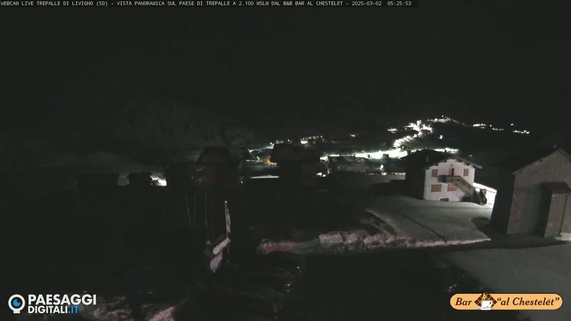 A nighttime, snow-covered village is illuminated by lights, viewed from a high vantage point.