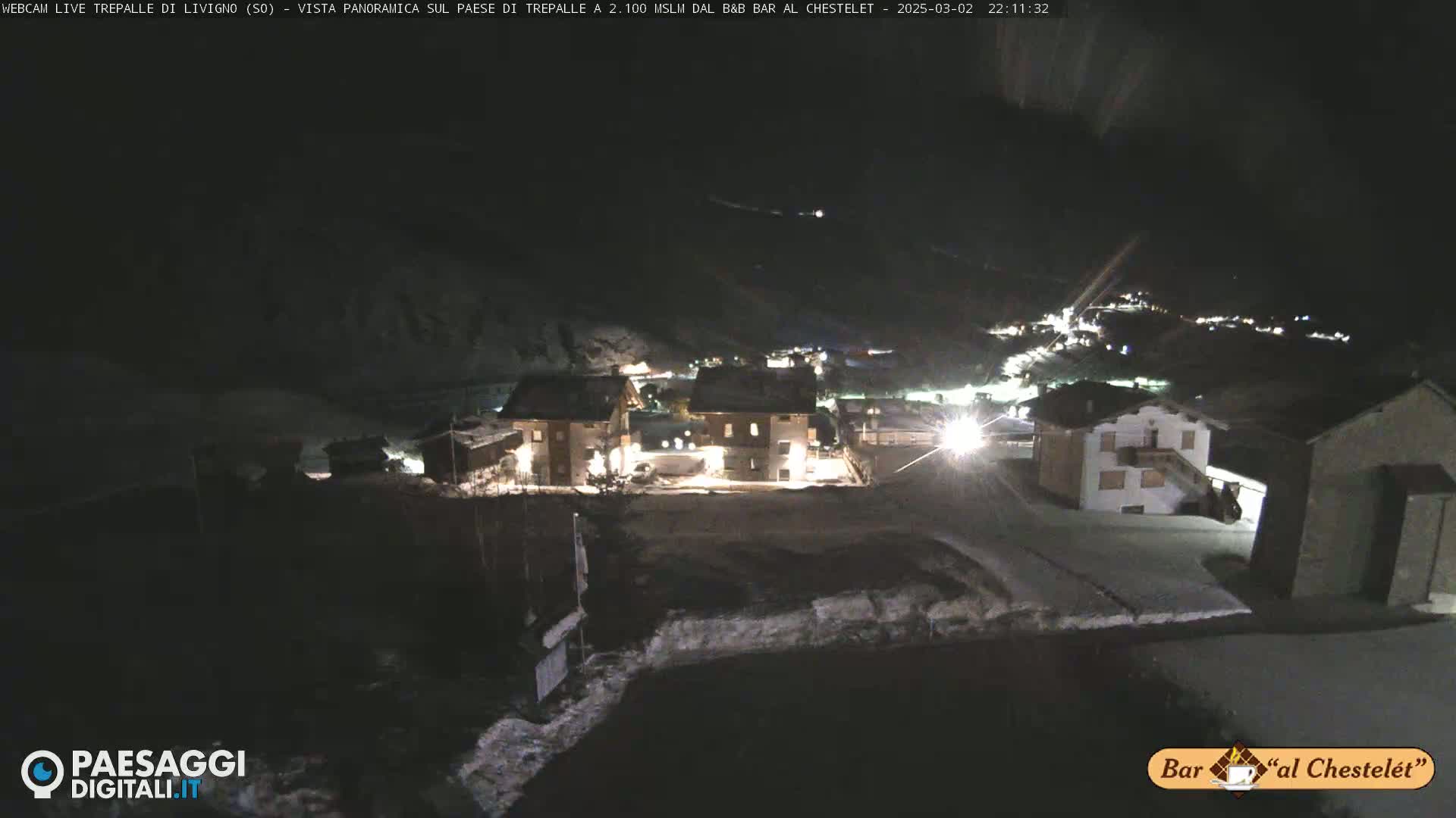 A nighttime, snow-covered village scene is illuminated by artificial lights, with a dark, mountainous background.