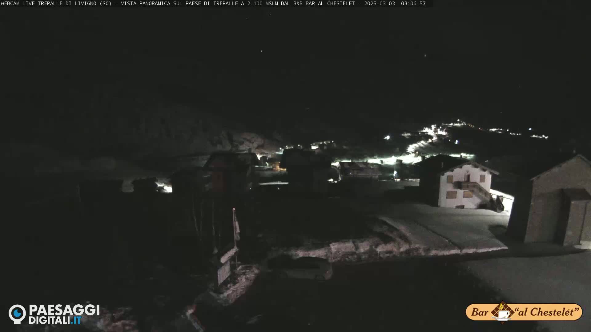A nighttime, snow-covered village is dimly lit, viewed from a high vantage point.
