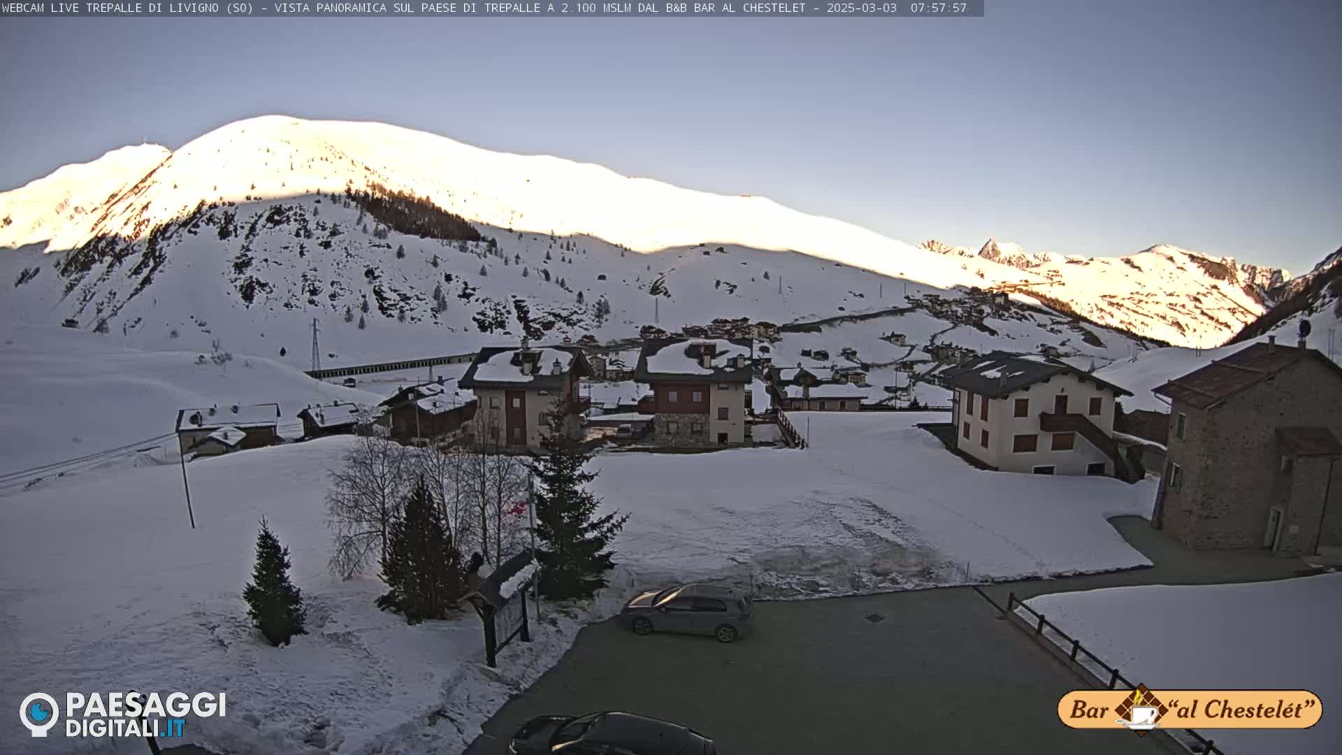 A snow-covered mountain village is bathed in the golden light of sunrise, with several buildings visible and a car parked in the foreground.