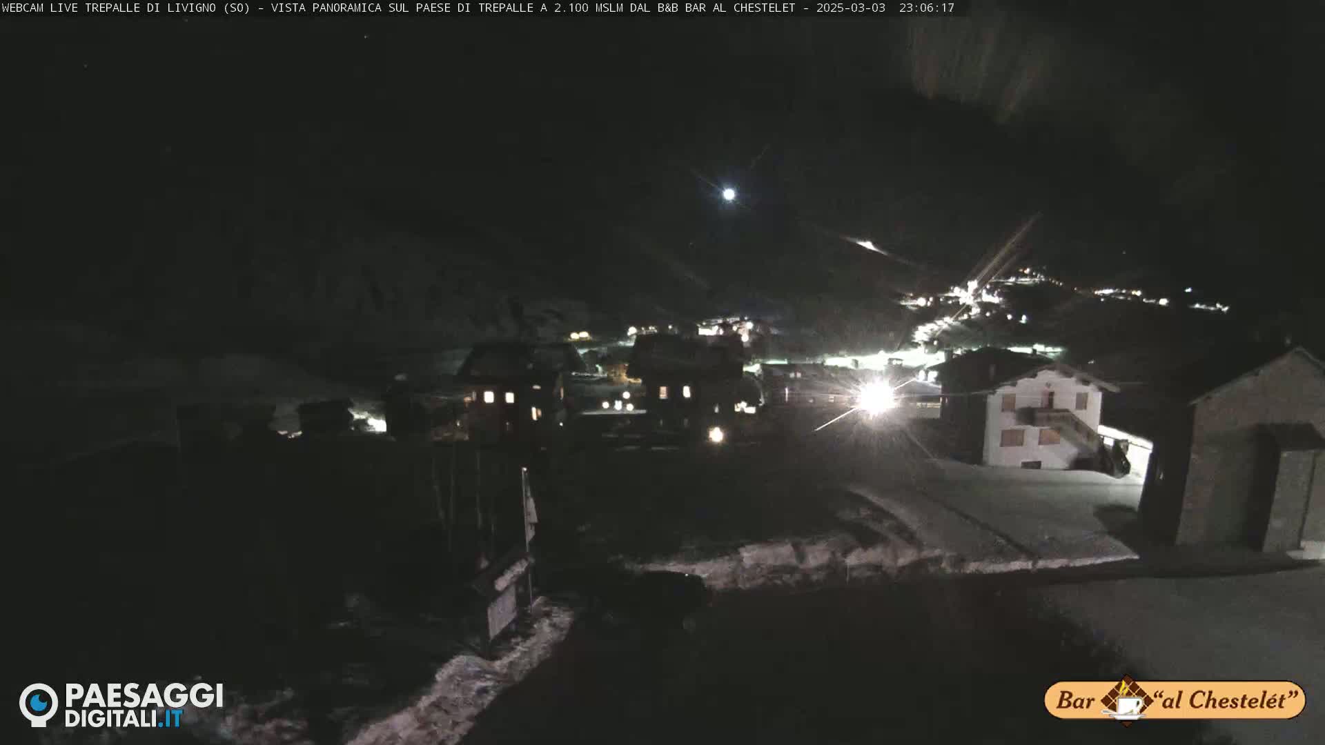A nighttime, snow-covered village is illuminated by lights, with a moon visible in the dark sky.