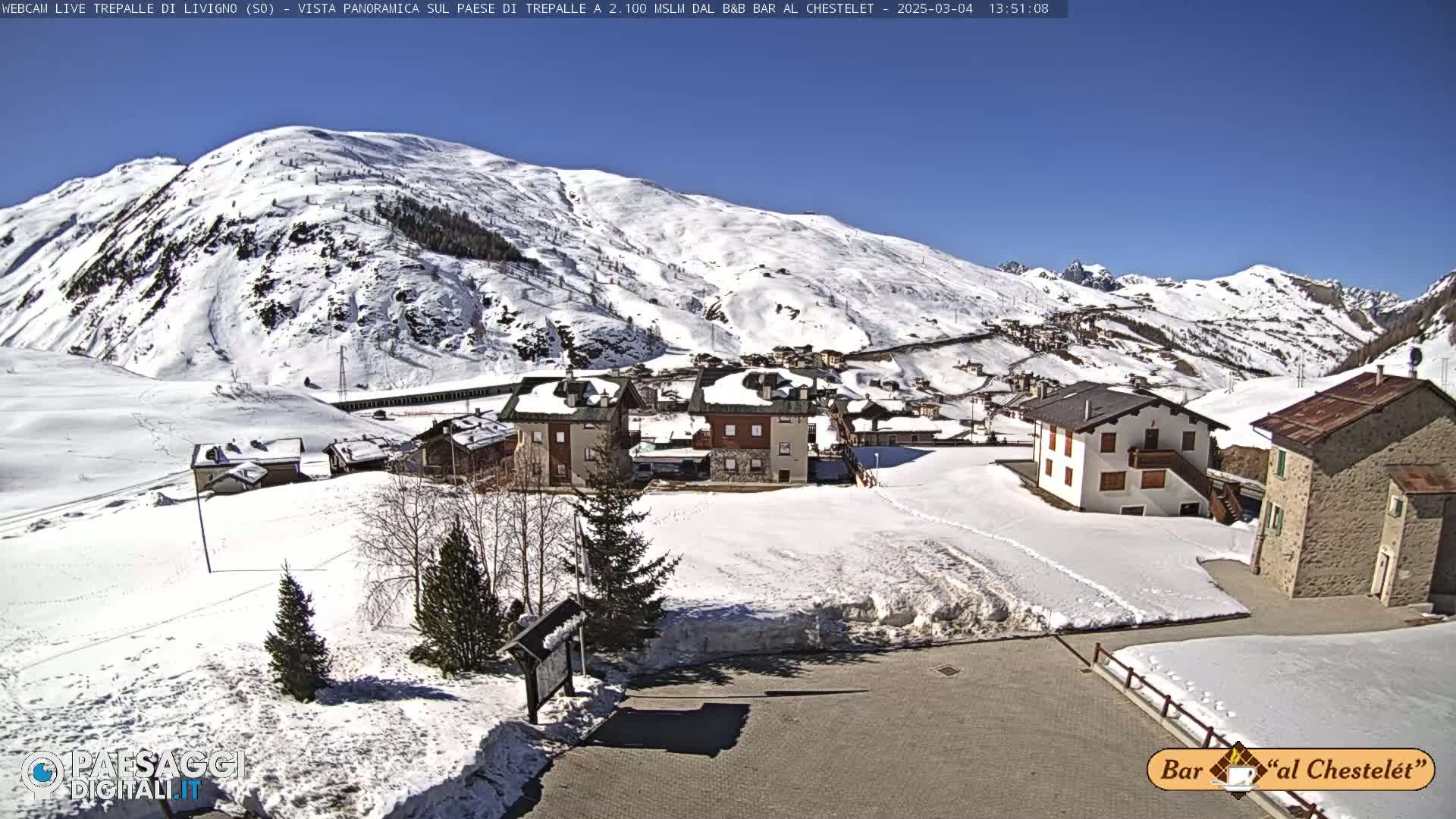 A snow-covered mountain village is seen on a sunny day, with surrounding hills also covered in snow.