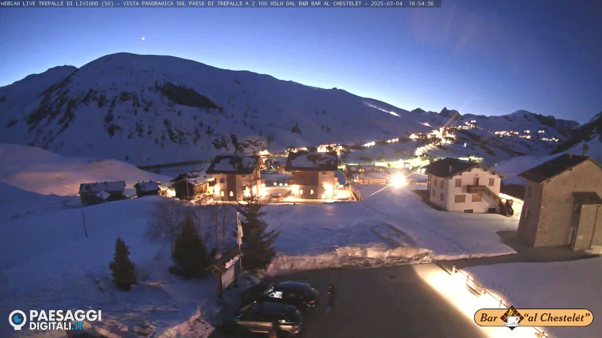 A snow-covered village at night is nestled in a valley between snow-capped mountains under a twilight sky.
