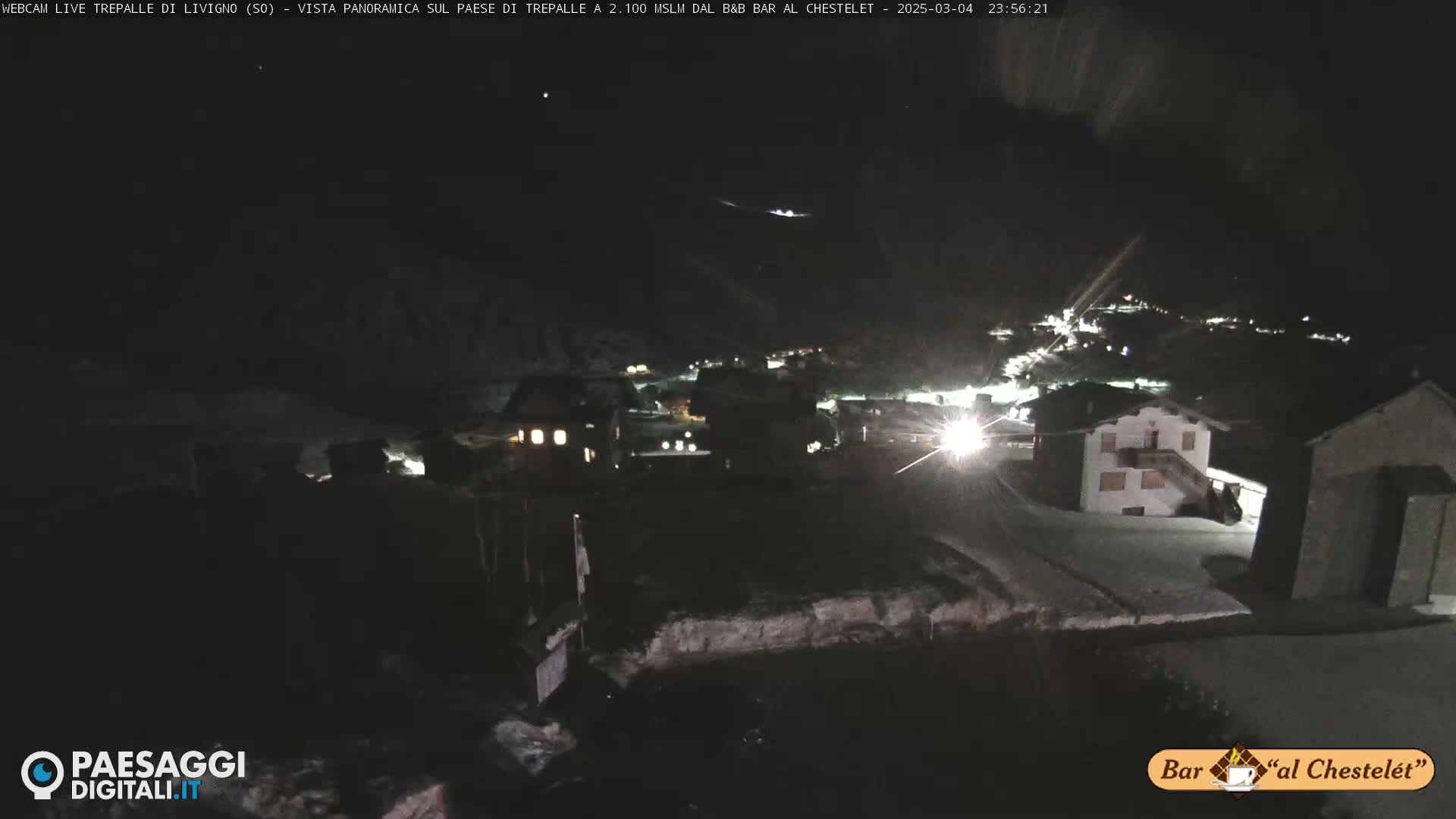 A nighttime, snow-covered village is dimly lit, with a few buildings visible under a dark sky.