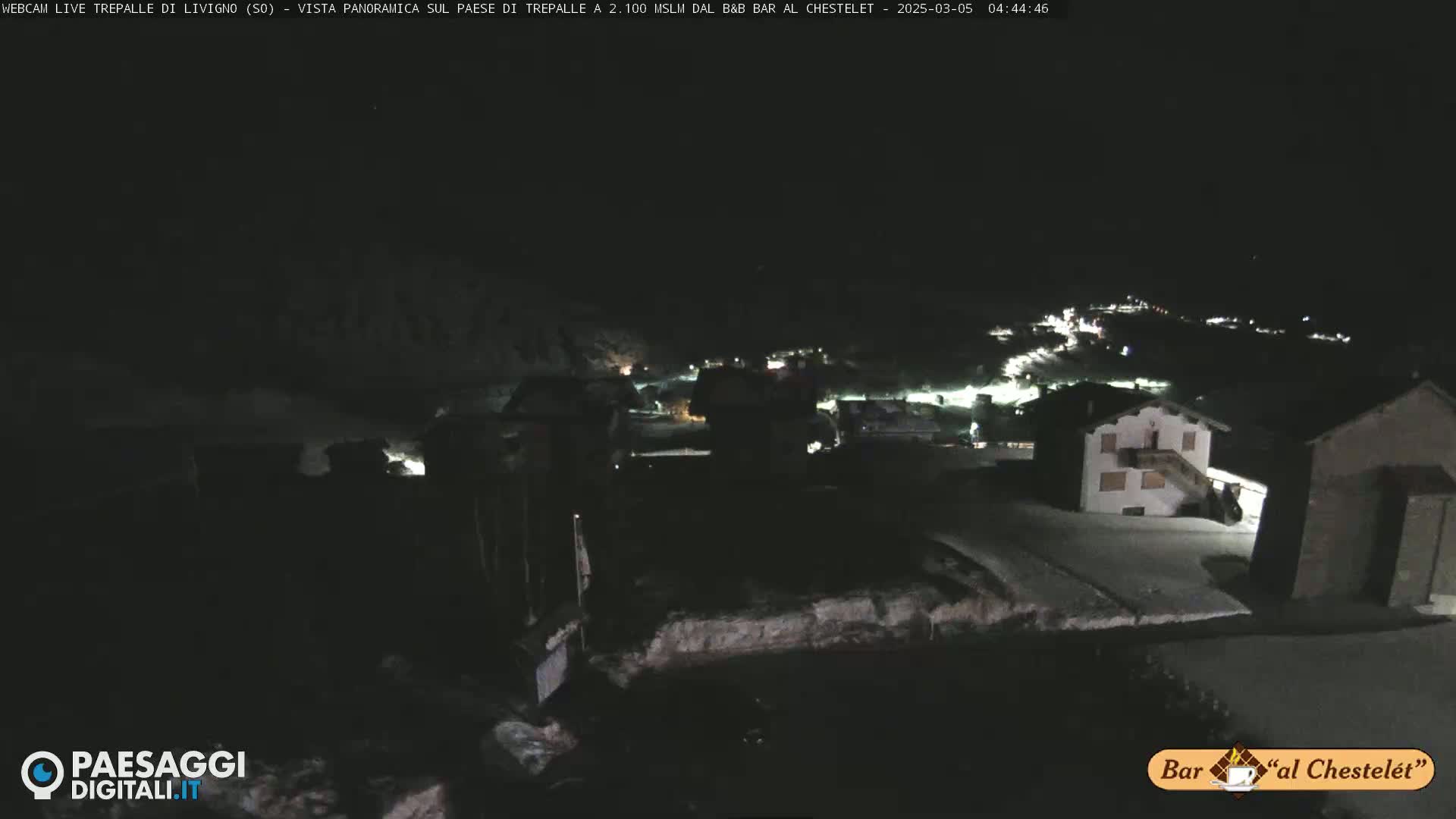 A nighttime, snow-covered village is visible from a high vantage point under a dark sky.