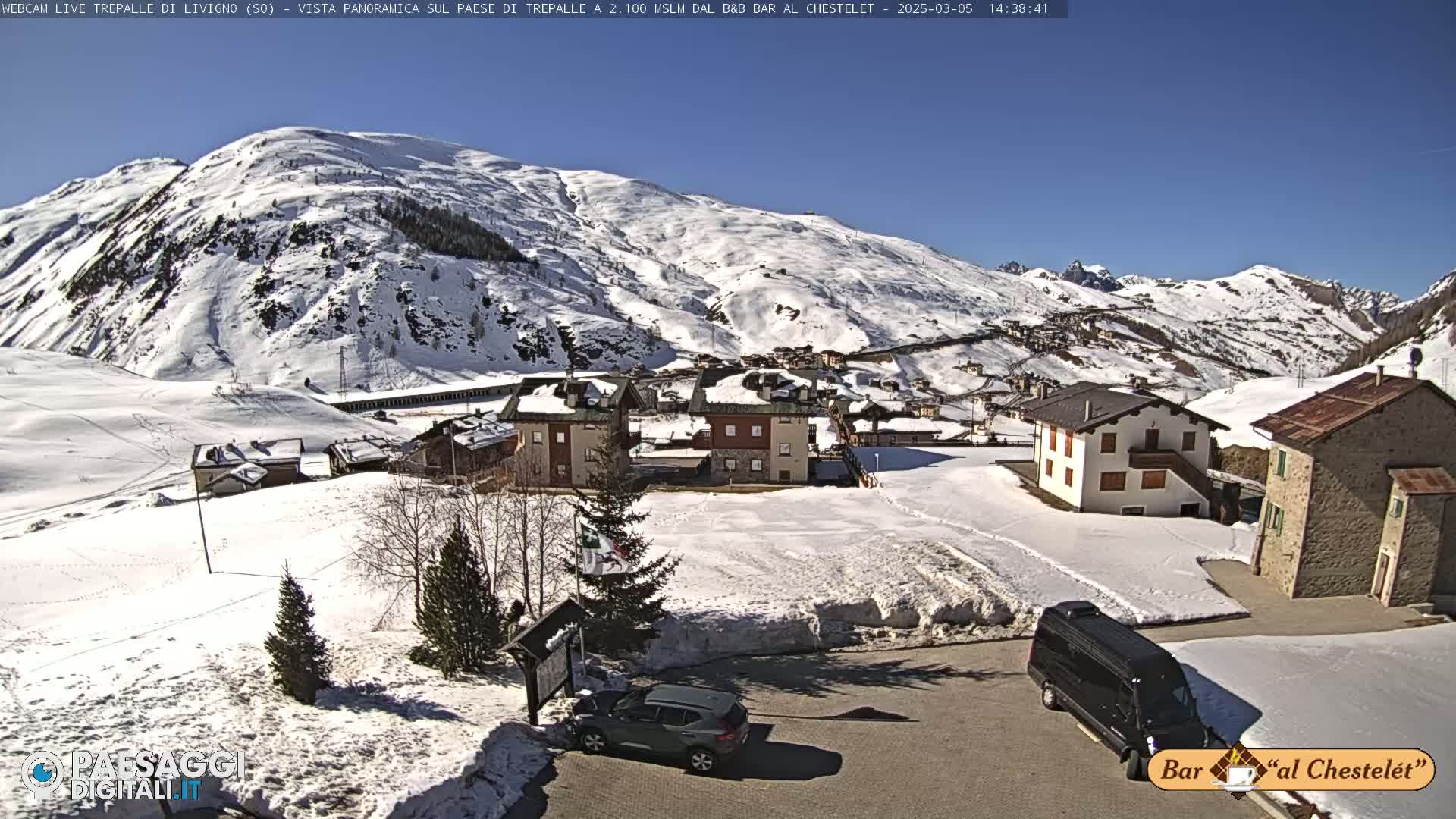 A sunny, snow-covered mountain village scene shows several buildings, a few vehicles, and a large snow-covered mountain in the background.
