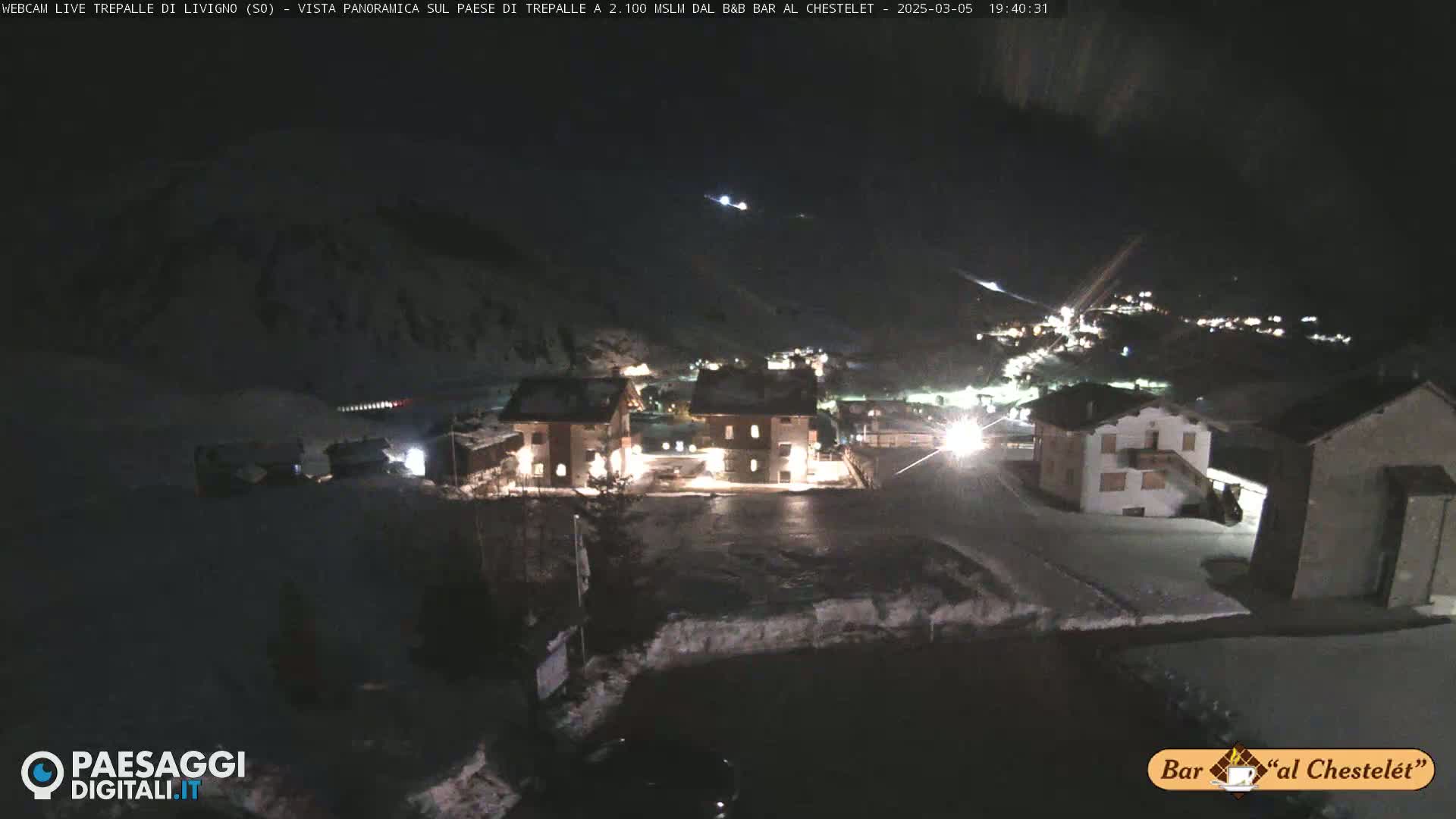 A nighttime, snow-covered village scene illuminated by building lights, nestled in a dark, mountainous area.