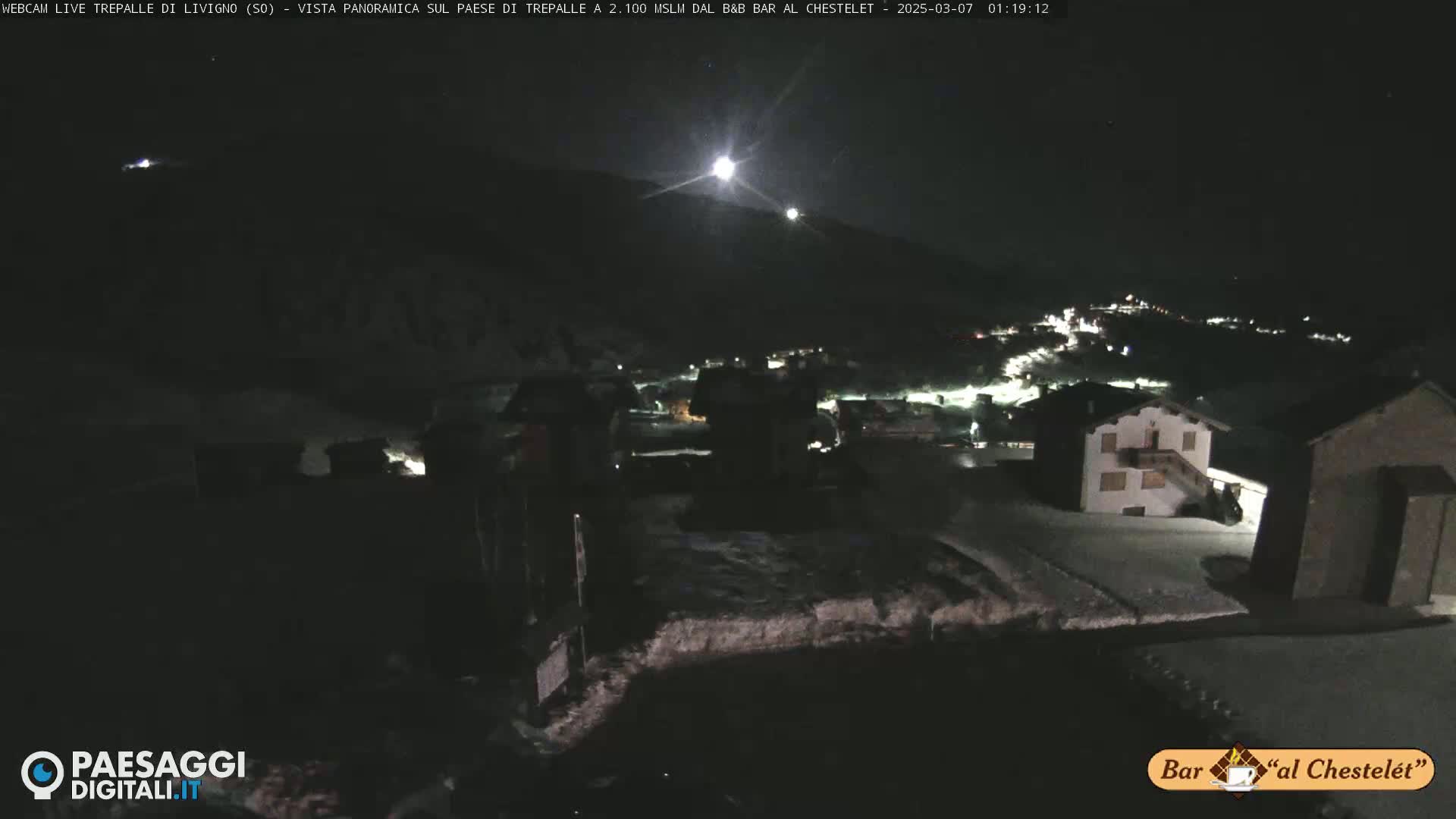 A nighttime, snow-covered village is illuminated under a bright moon and clear sky.