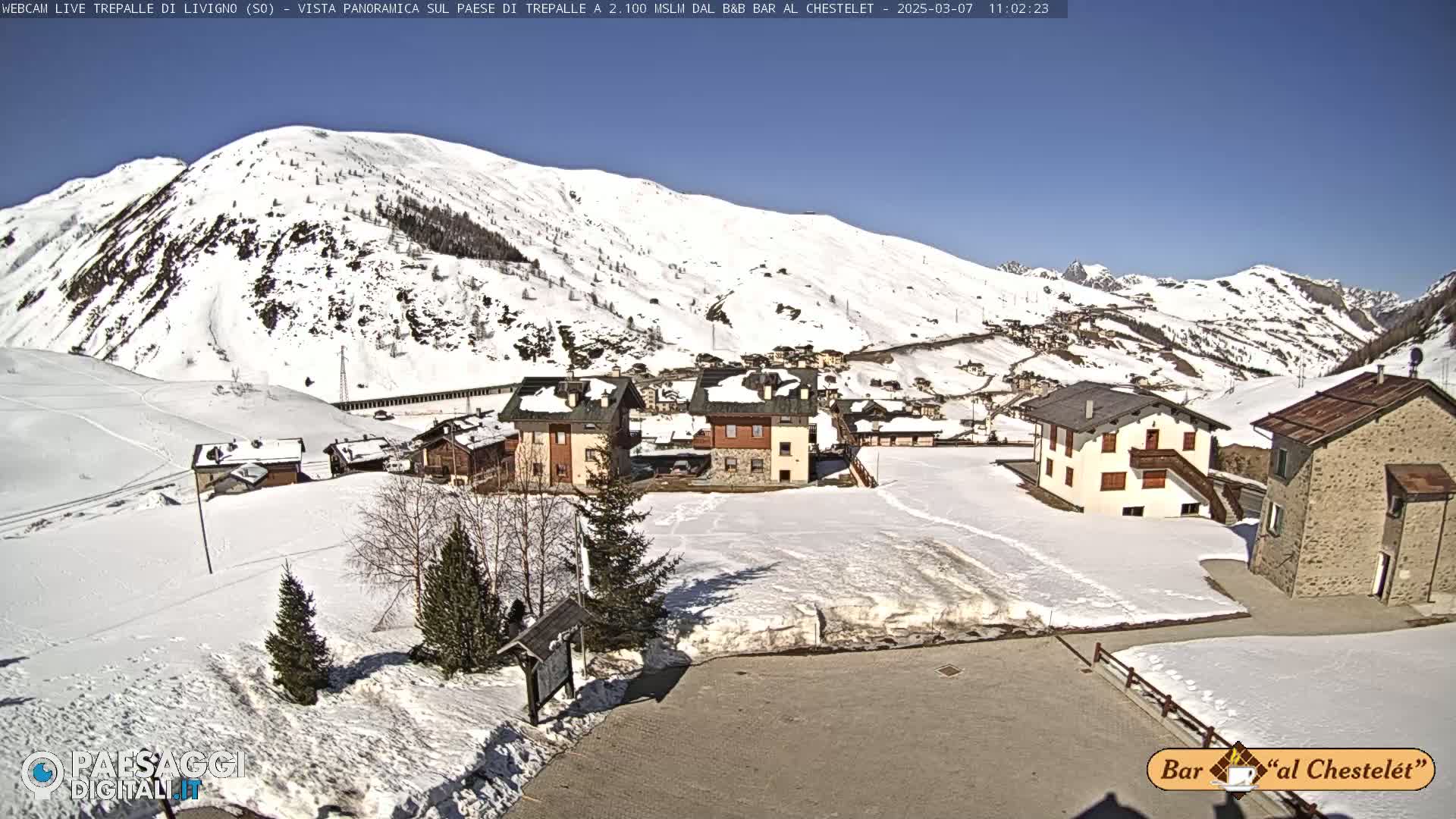 A snow-covered village nestled in a valley surrounded by snow-capped mountains under a clear, sunny sky.