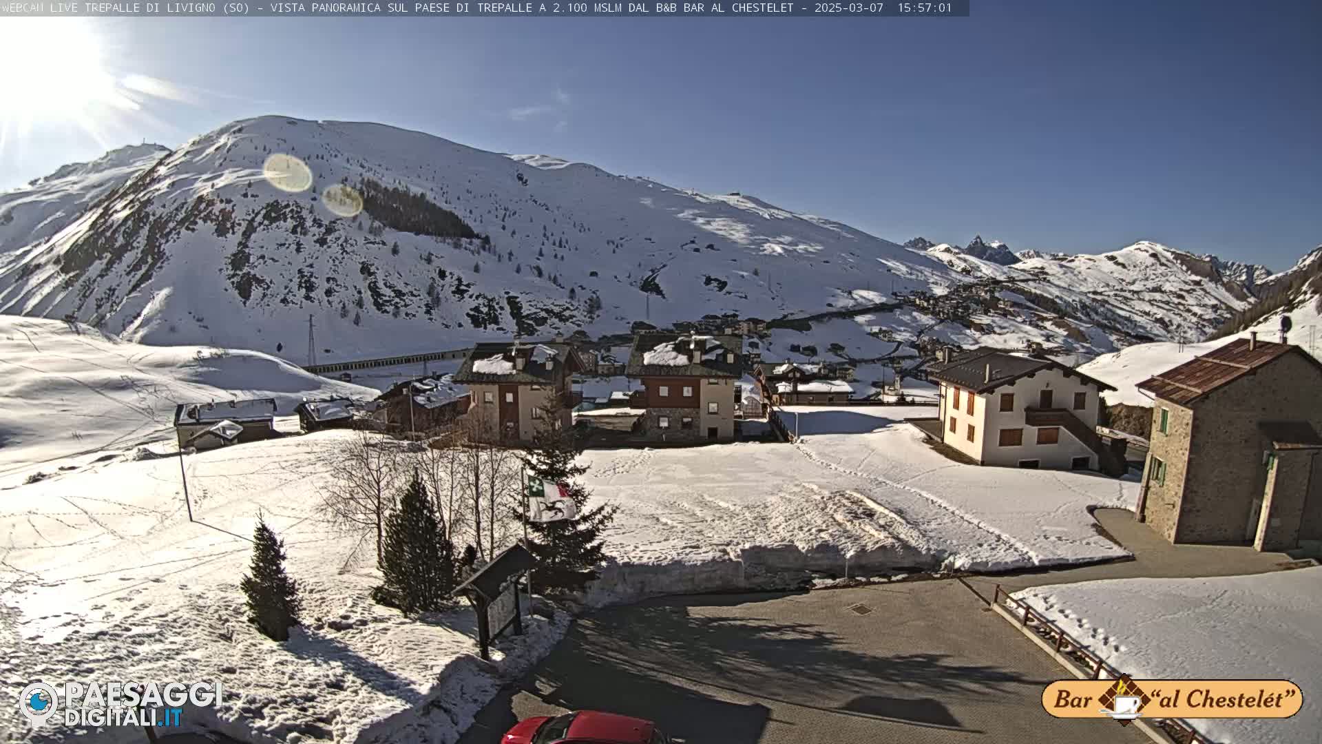 A snow-covered mountain village is nestled at the base of a sunlit, snow-dusted mountain range under a clear blue sky.