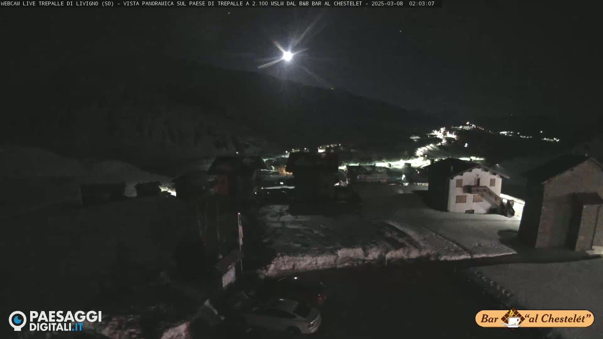 A nighttime, snow-covered village is illuminated by a bright moon under a clear sky.