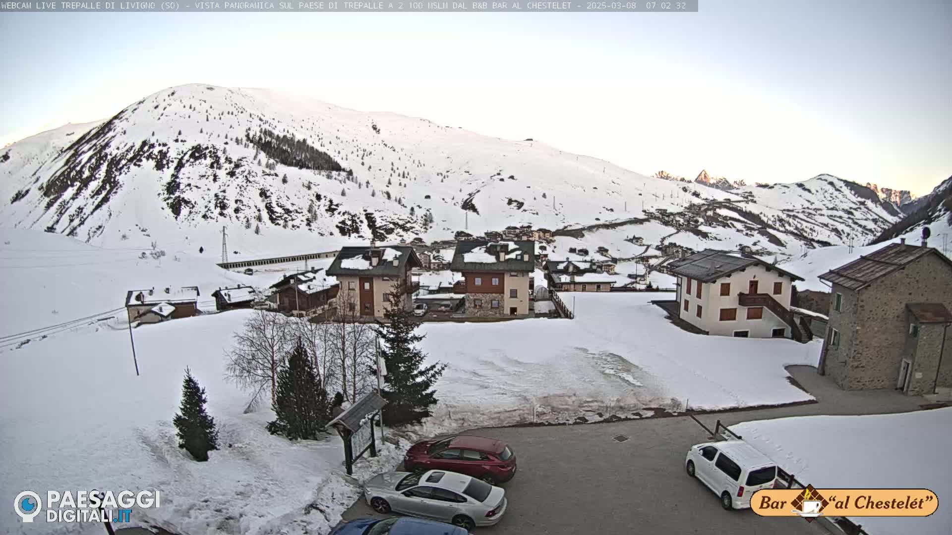 A snow-covered mountain village with several buildings and a few cars parked in a lot, under a clear, bright sky.