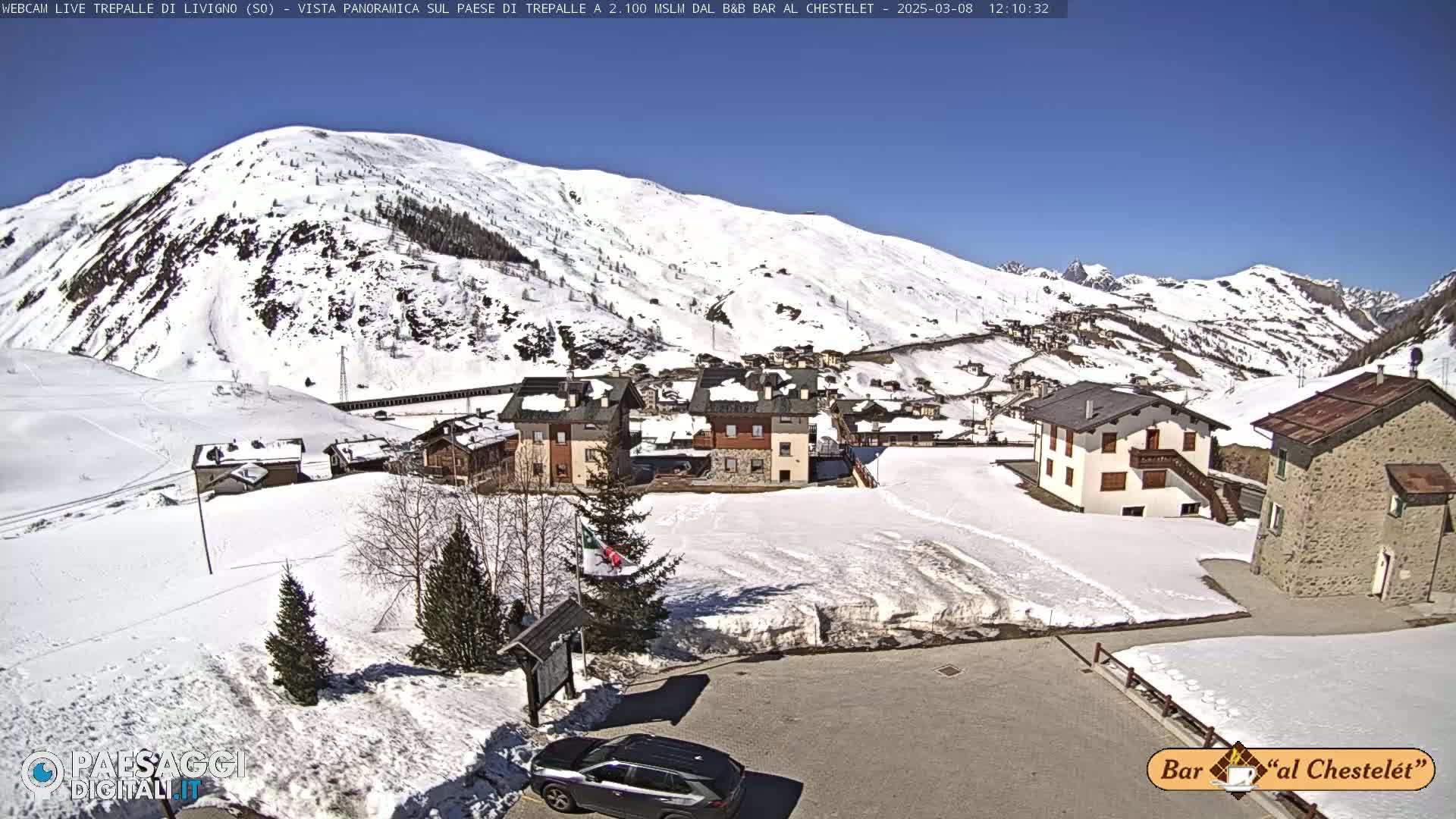 A sunny, snow-covered mountain village scene with several buildings and a car parked in a lot.
