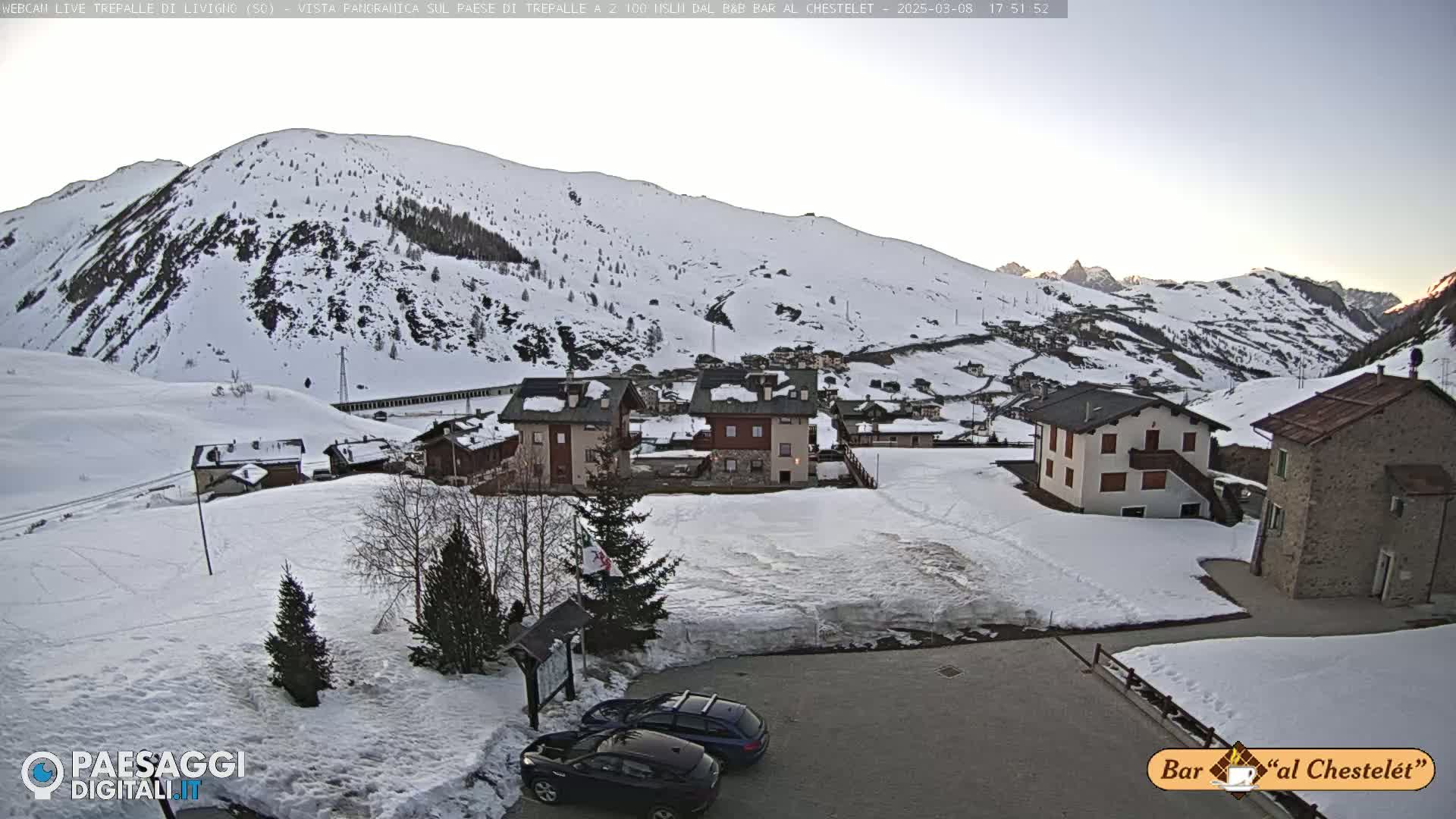 A snow-covered village nestled in a mountain valley at dusk, with two cars parked in a small lot.