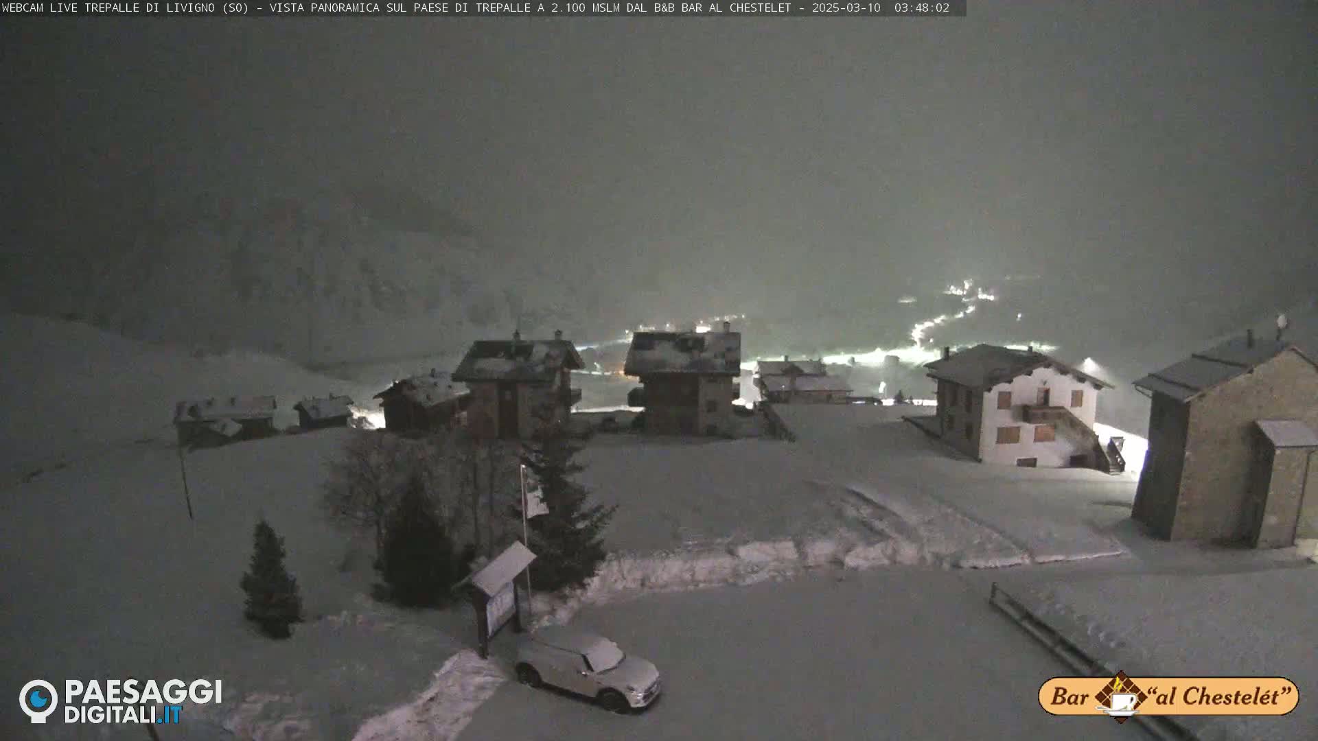 A snow-covered village at night is illuminated by scattered lights, with snow falling in the dark, overcast sky.