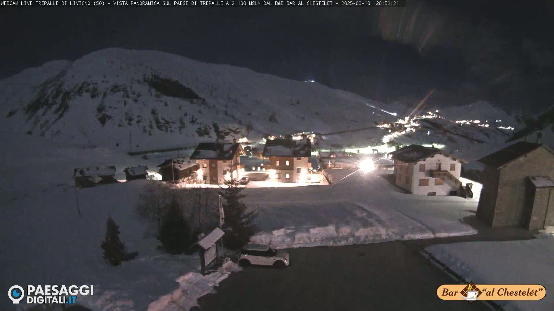 A snow-covered village at night, illuminated by lights, is nestled in a mountain valley under a dark sky.