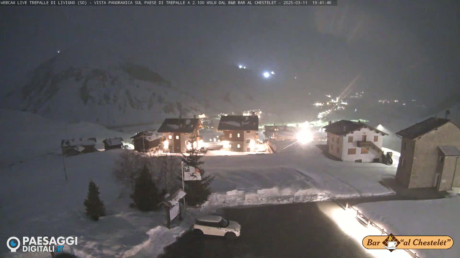 A snow-covered village at night is illuminated, with lightly falling snow and mountains in the background.