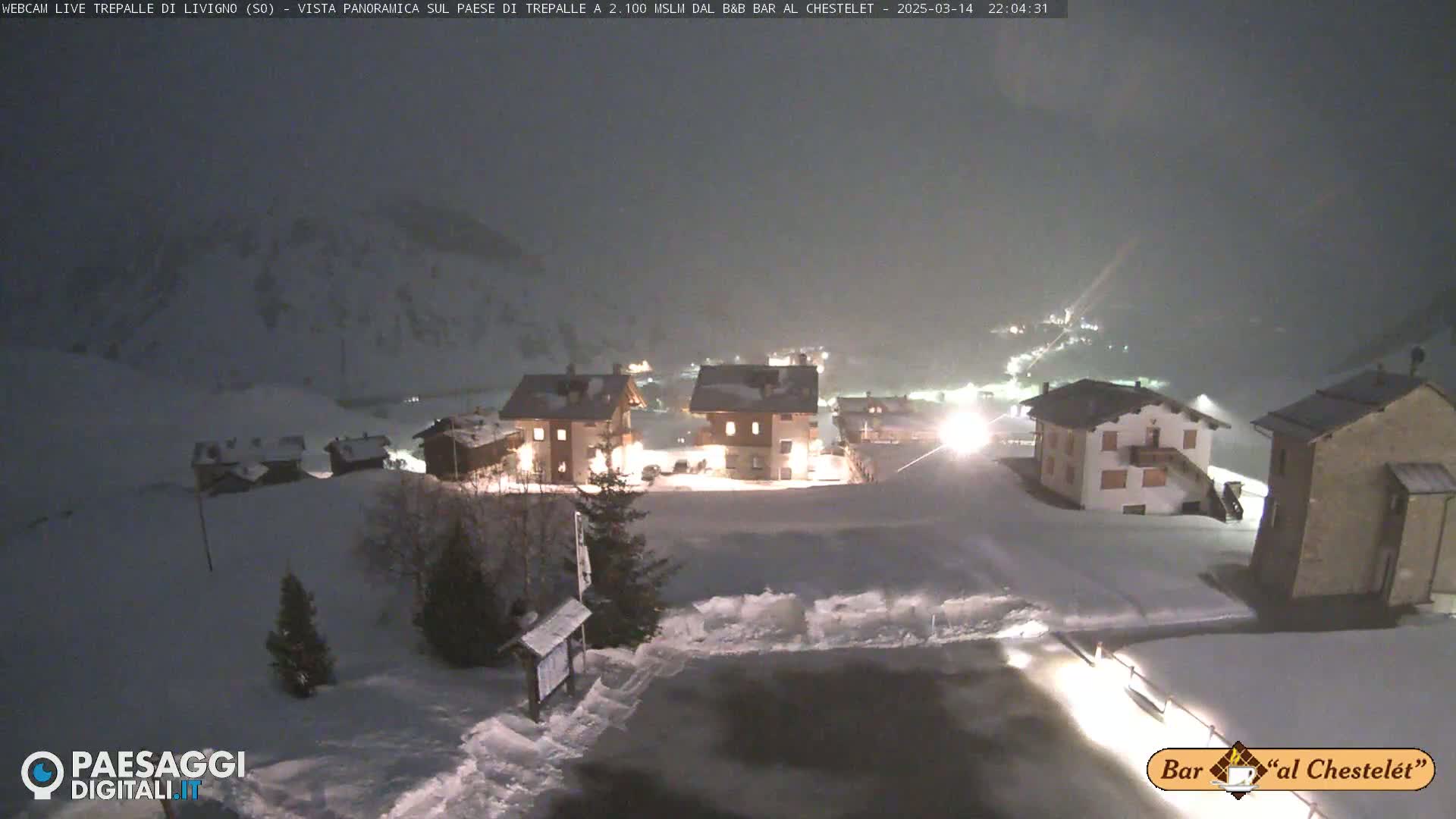 A snow-covered village is illuminated at night under a dark, snowy sky.
