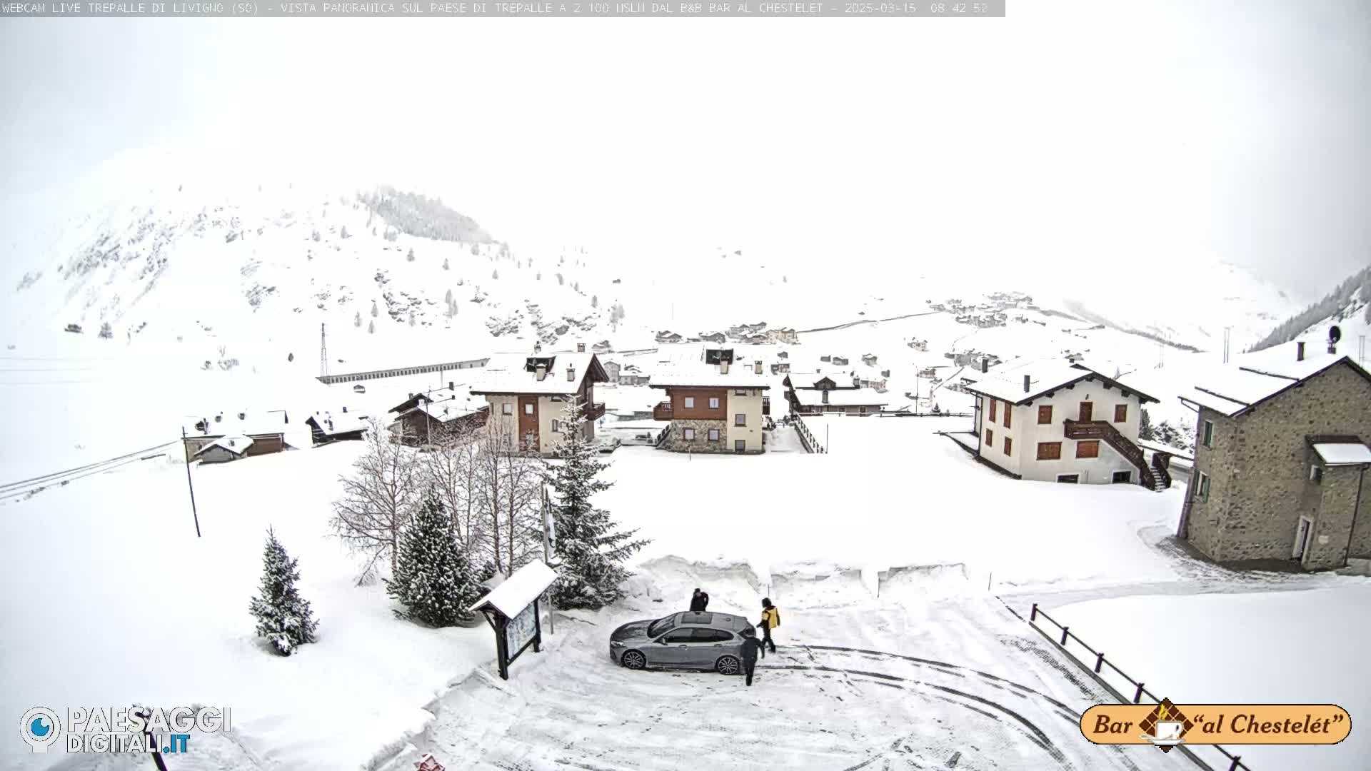 A snow-covered village nestled in a mountainous region, with a car and several people in a parking area.