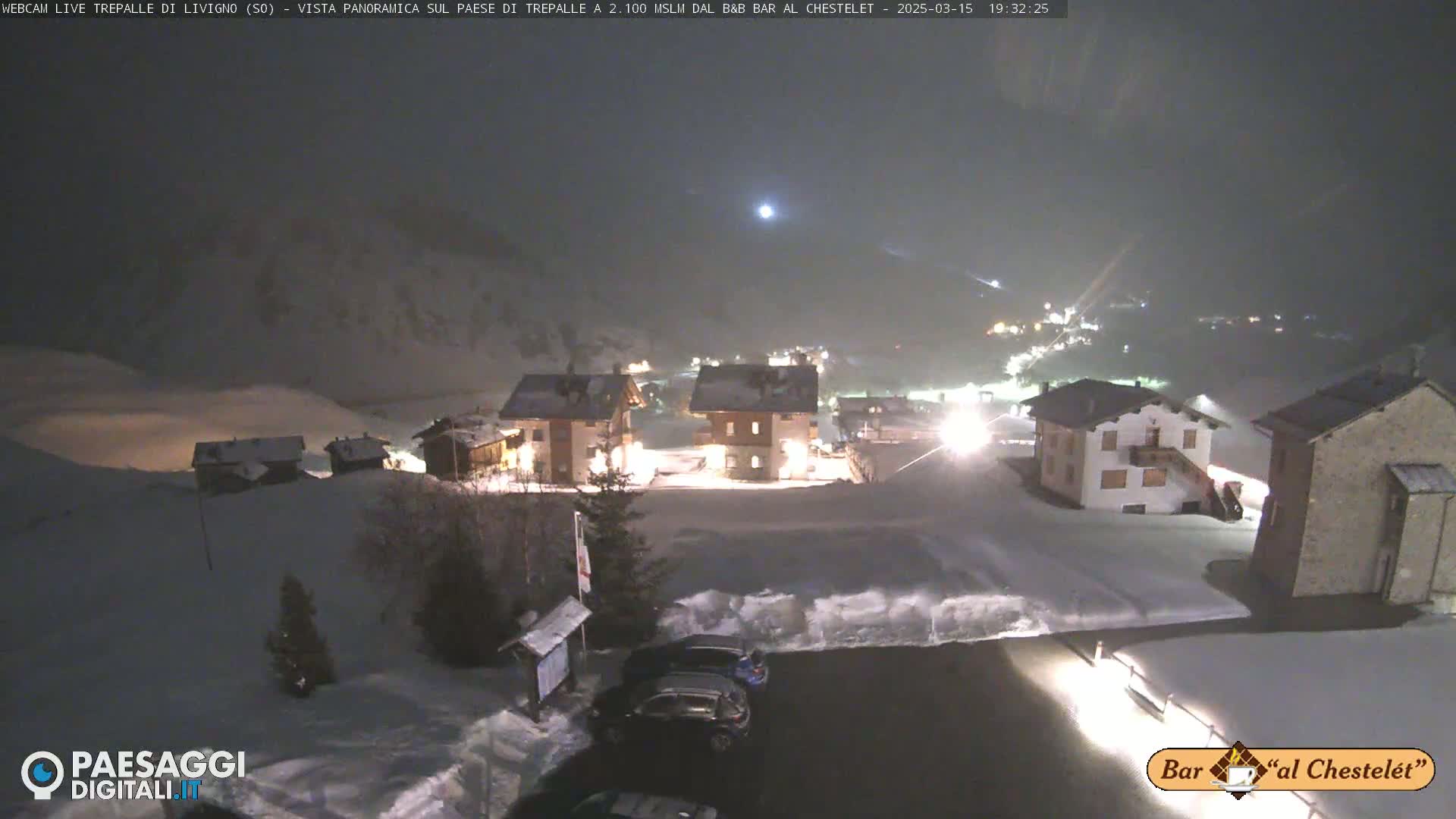 A snow-covered village at night is illuminated, with a few cars parked in the foreground under a hazy, dark sky.