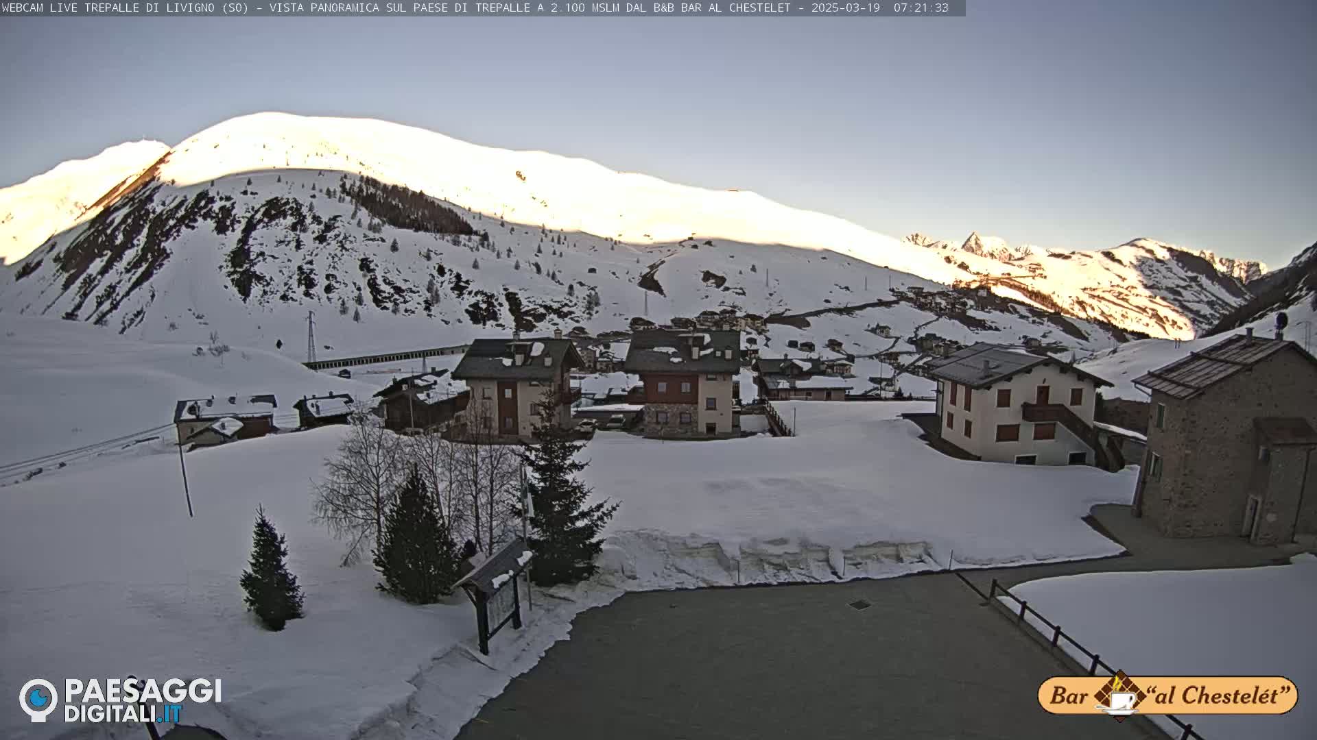 A snow-covered village nestled in a valley between snow-capped mountains under a clear, sunrise sky.