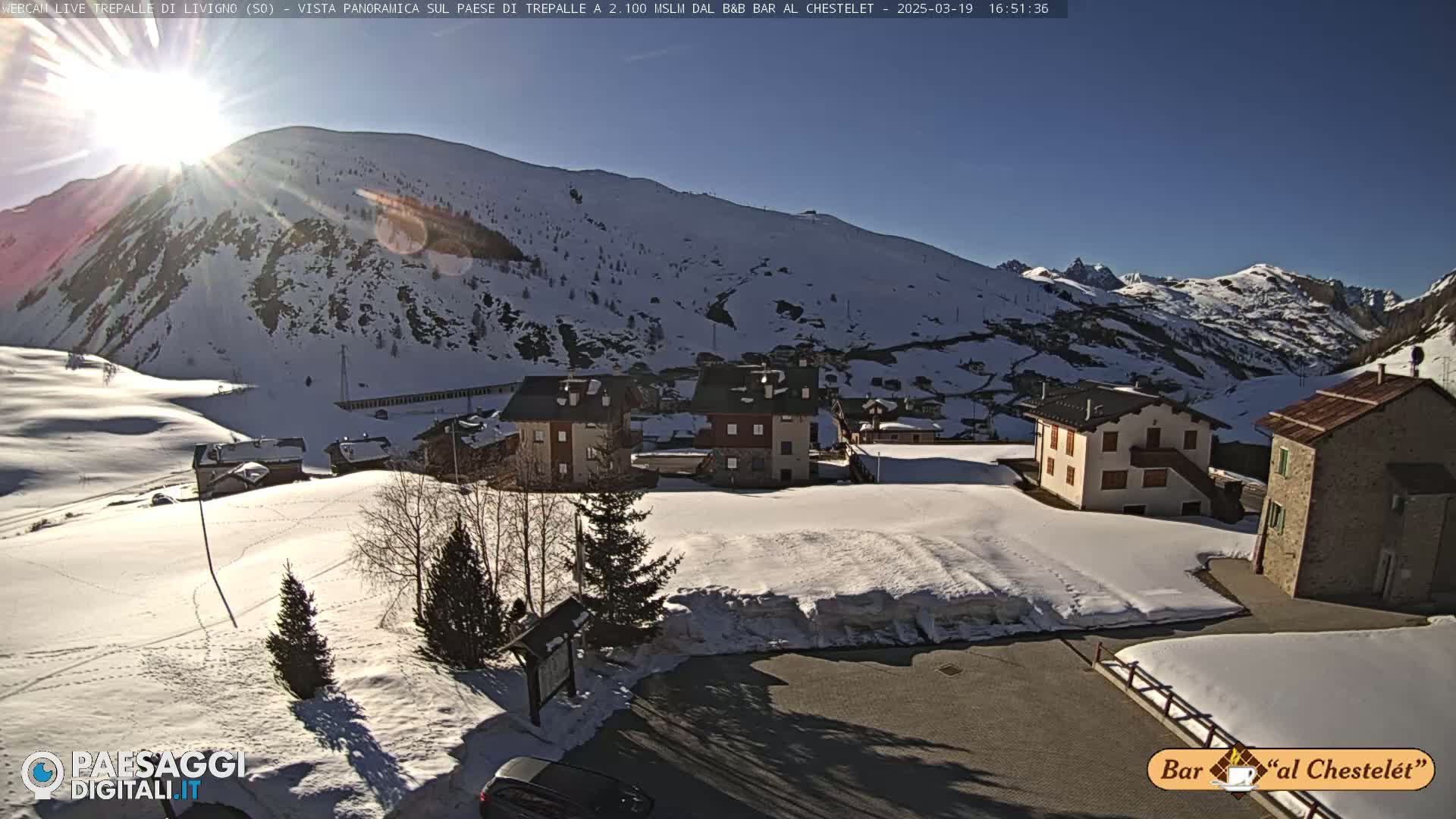 A sunny, snow-covered mountain village with several buildings is seen from an elevated vantage point.