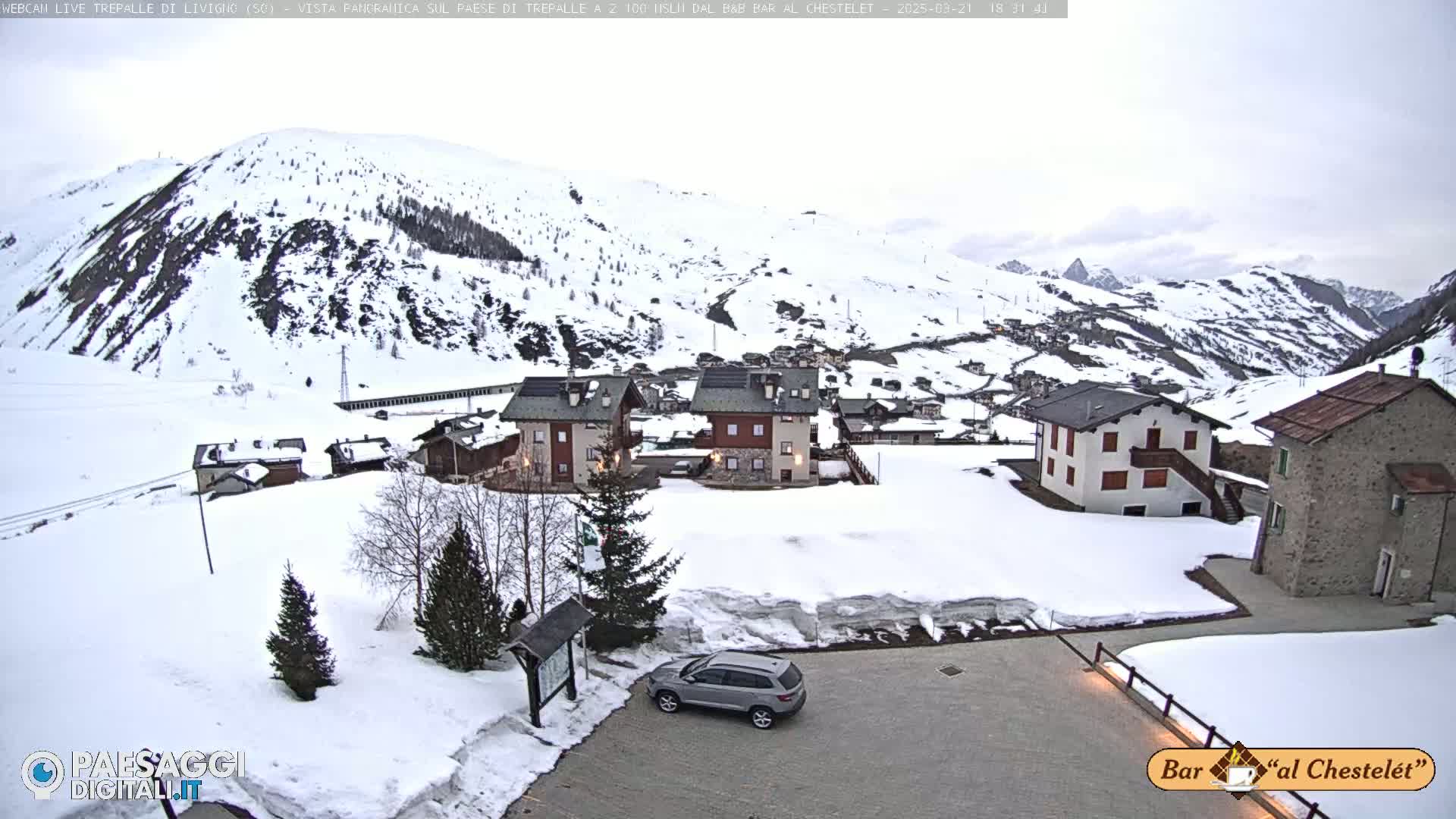 A snowy mountain village scene shows several buildings, a single car parked in a snowy lot, and a background of snow-covered mountains under an overcast sky.
