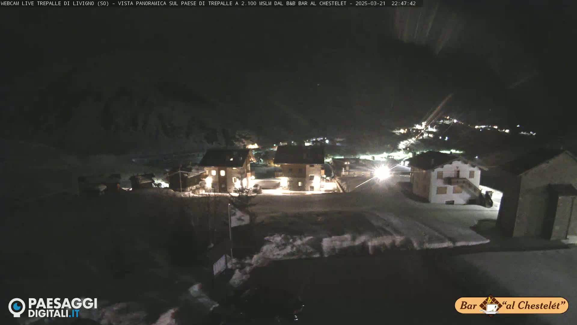 A nighttime, snowy scene shows a small village with several buildings illuminated, under a dark sky.