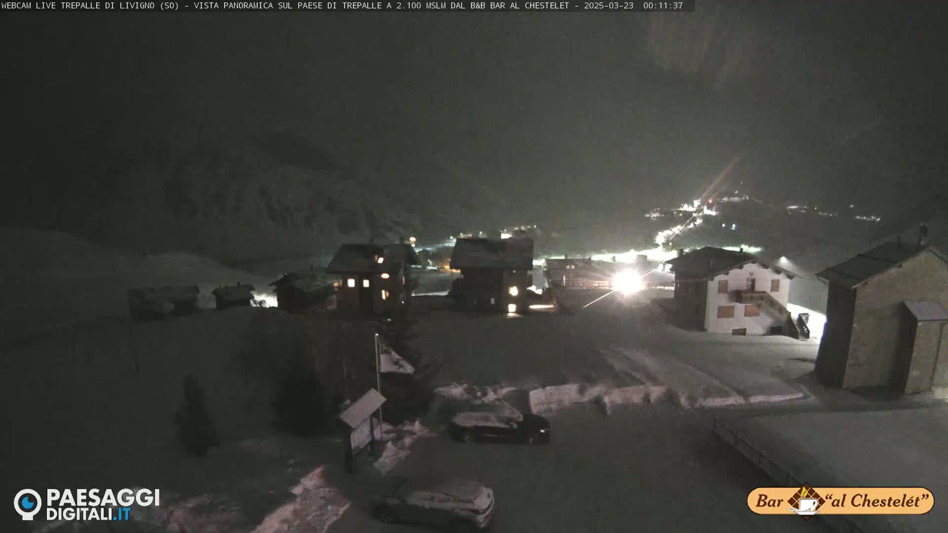 A snow-covered village at night is illuminated by scattered lights, with some cars parked in a lot.