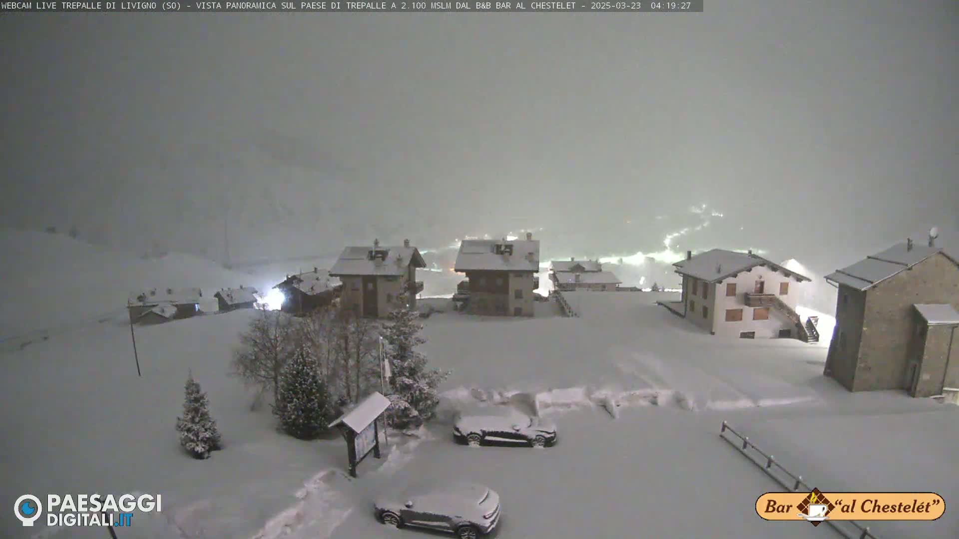 A snow-covered village scene at night, with several buildings illuminated and cars parked in the snow.
