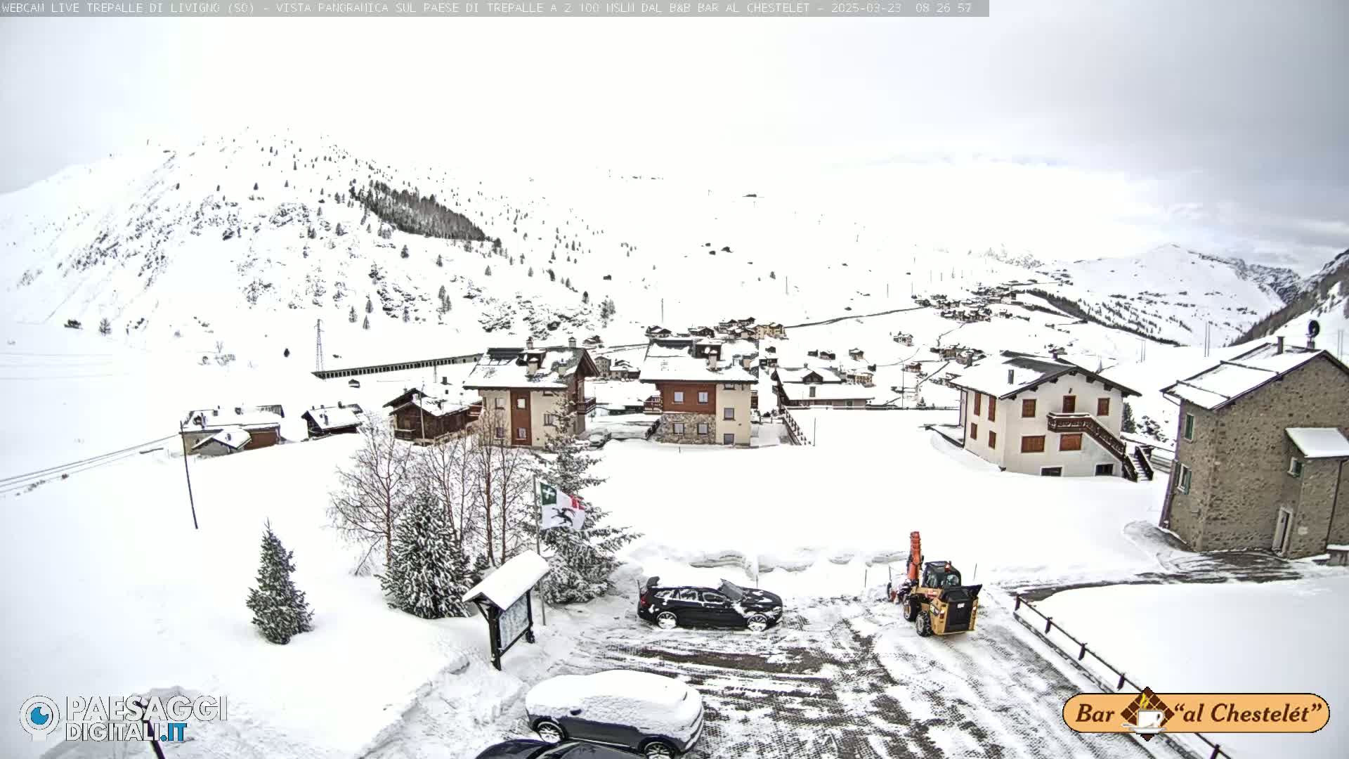 A snow-covered village nestled in a mountainous region under an overcast sky, with several cars parked in a snow-laden parking lot and a snow plow clearing a road.
