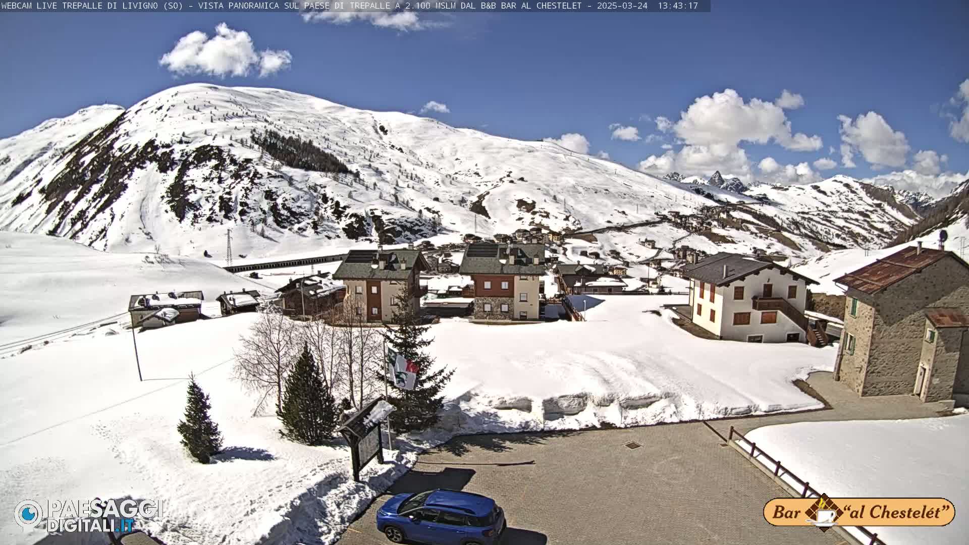 A snow-covered village nestled in a mountain valley under a mostly sunny sky with some scattered clouds.