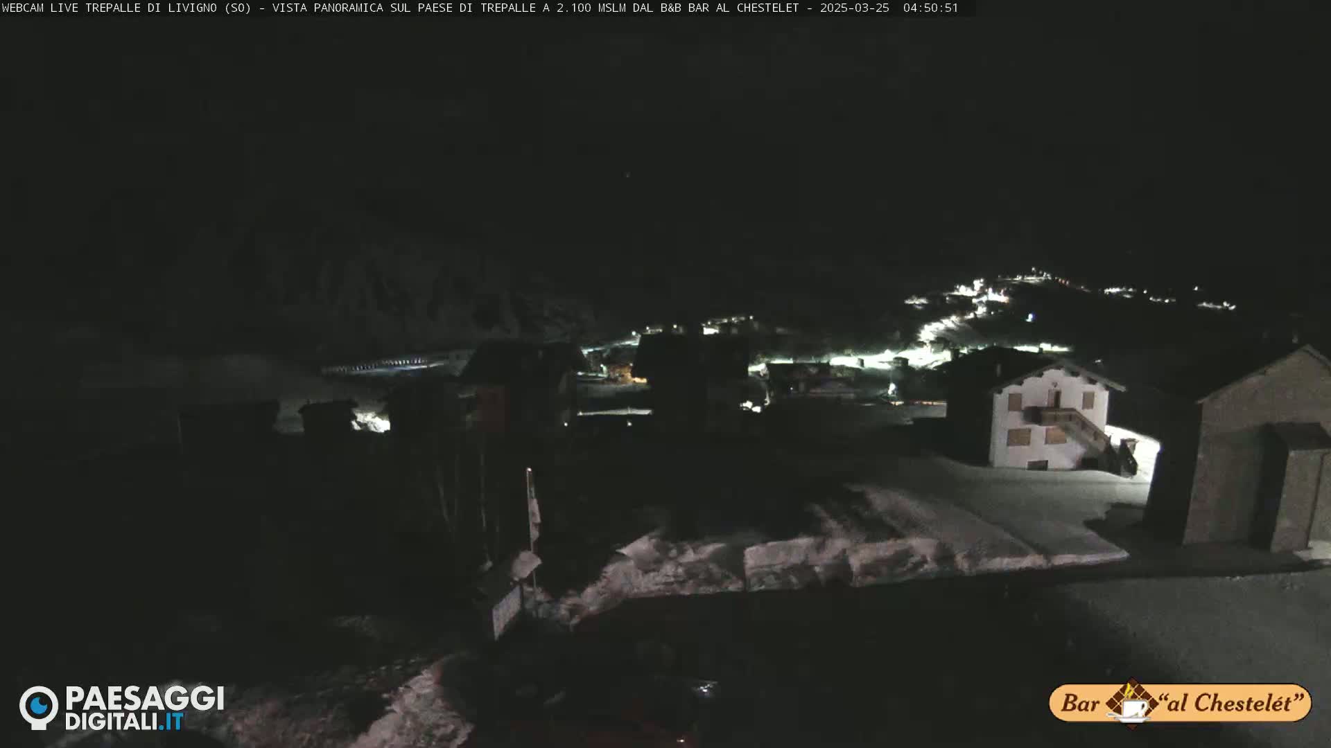 A nighttime, snowy scene shows a small village with dimly lit buildings under a dark sky.