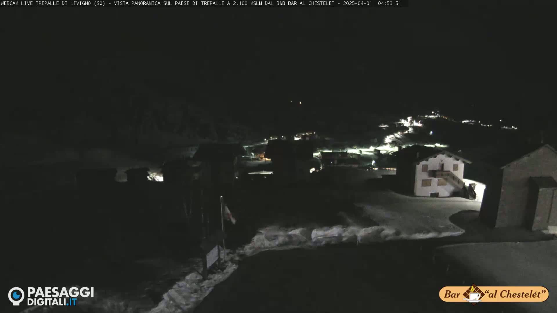 A nighttime, snow-covered village is illuminated by scattered lights under a dark sky.