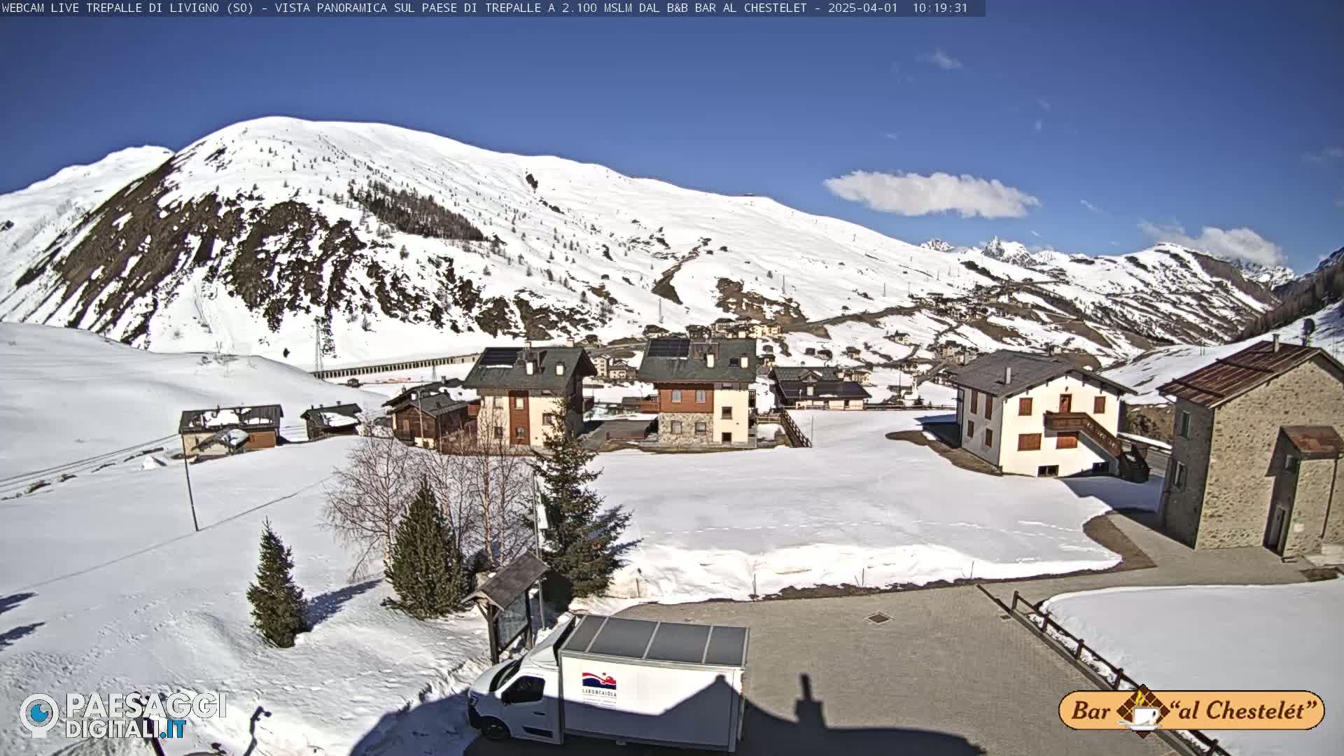 A sunny, snow-covered mountain village scene shows several buildings nestled amongst patches of snow, with a larger snow-covered mountain in the background.