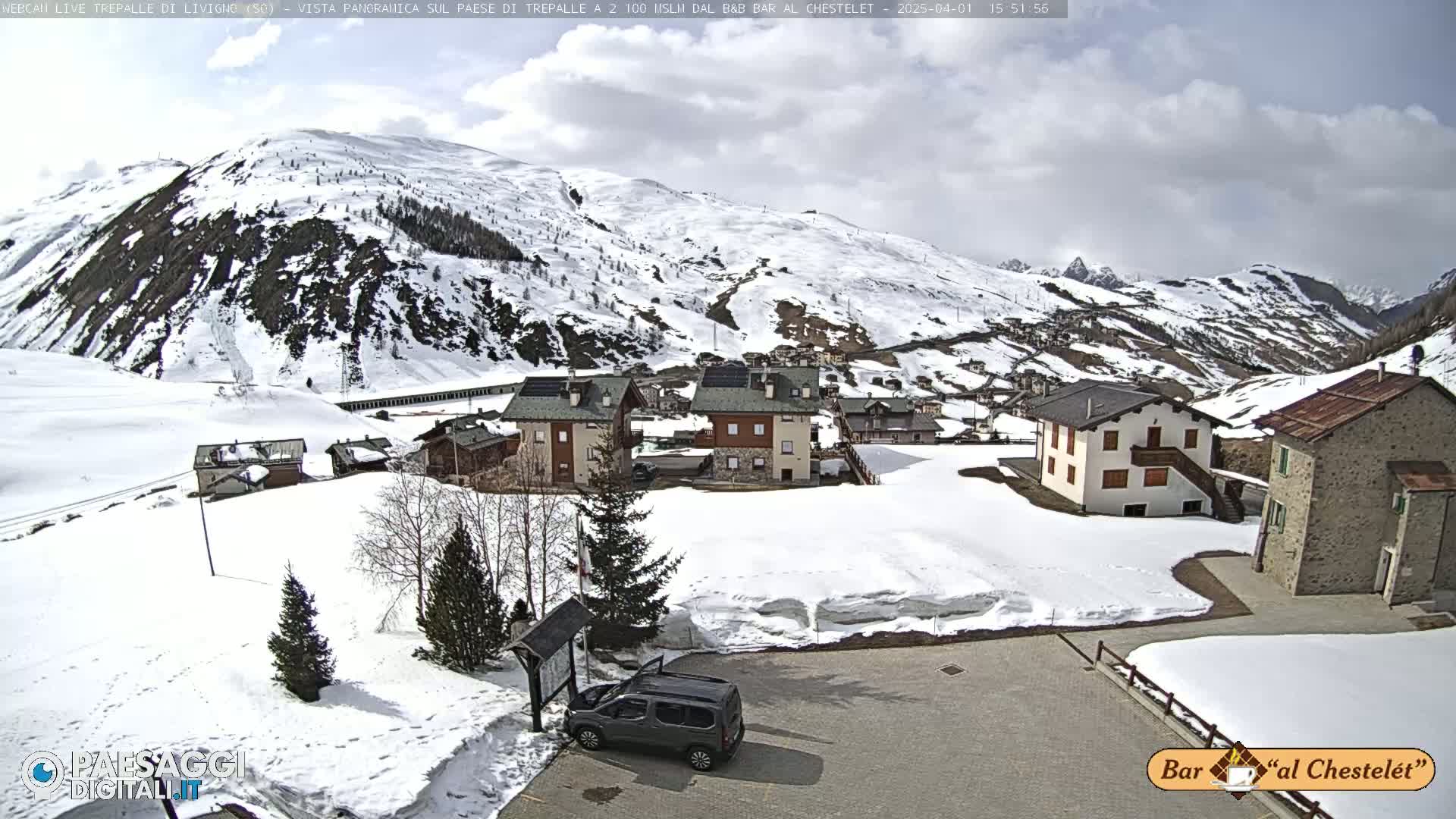 A snow-covered village nestled in a valley surrounded by snow-dusted mountains under a partly cloudy sky.
