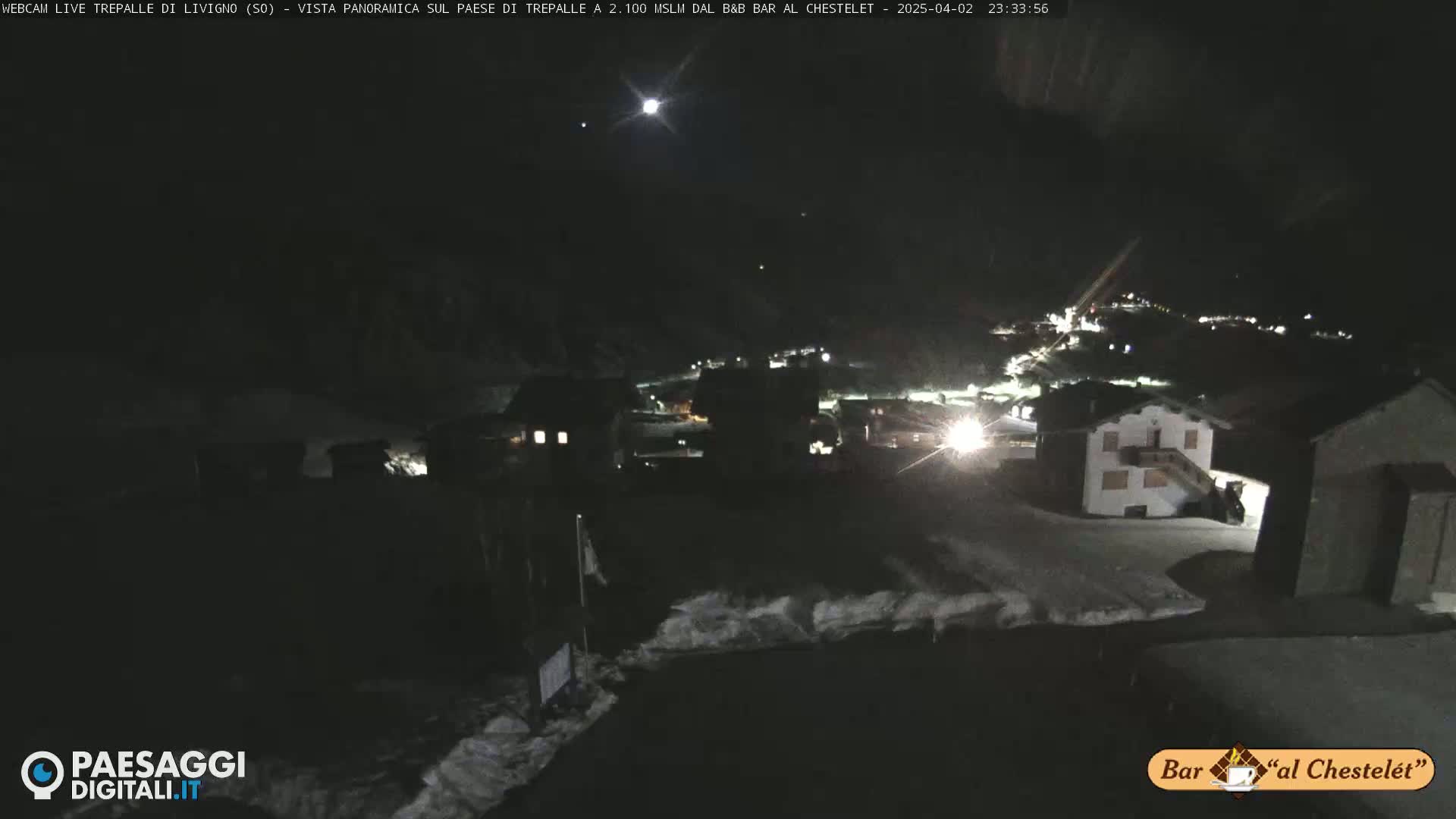 A nighttime, snow-covered village scene is illuminated by scattered lights under a moonlit sky.