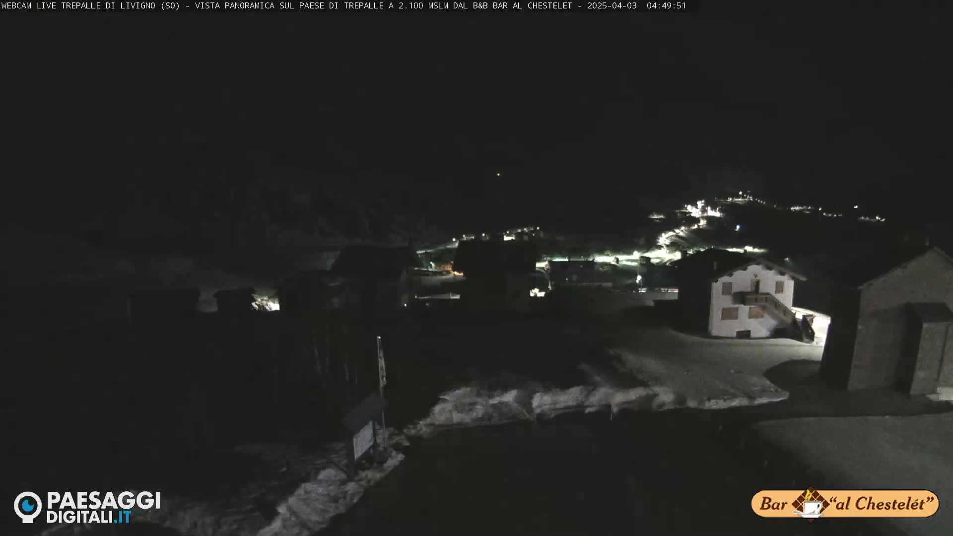 A nighttime, snow-covered village is dimly lit, with a few buildings and lights visible in the distance under a dark sky.