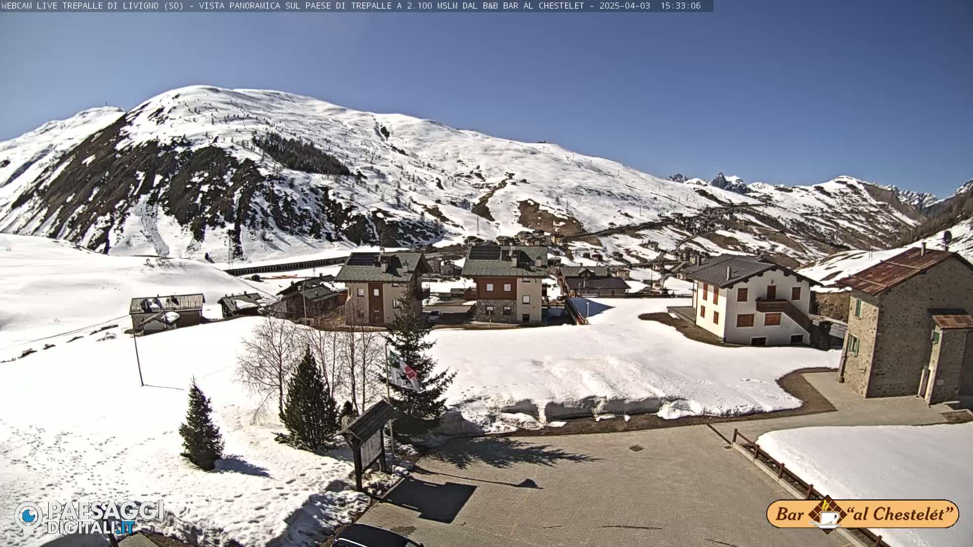 A sunny day reveals a snow-covered mountain village nestled below a snow-capped mountain range.