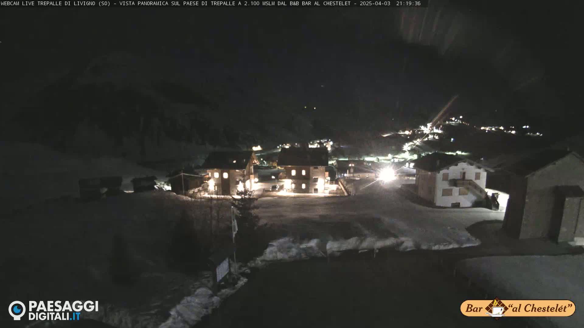 A nighttime, snow-covered village is illuminated by lights, with dark mountains in the background.