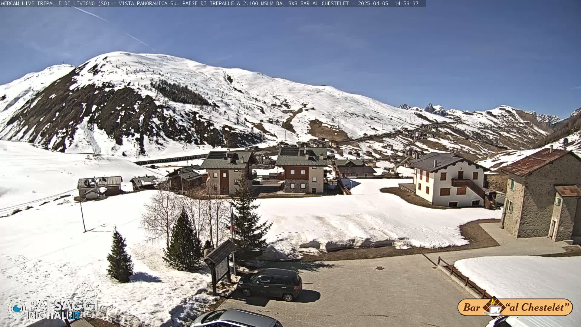 A sunny day reveals a snow-covered mountainside village with several buildings, some cars parked, and patches of snow remaining on the ground.