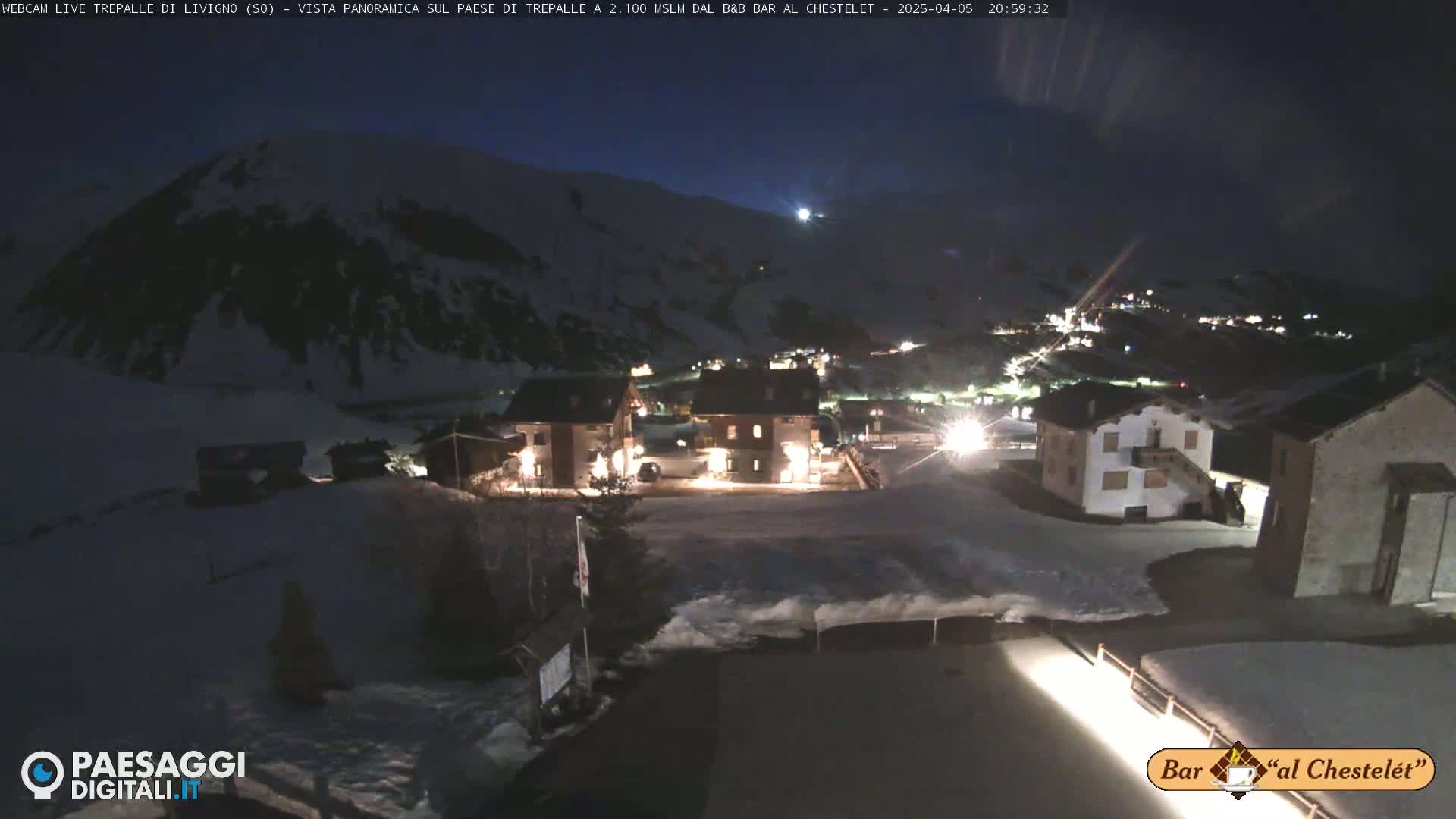 A nighttime, snow-covered village is illuminated, nestled below a dark, snow-dusted mountain under a clear, dark sky.
