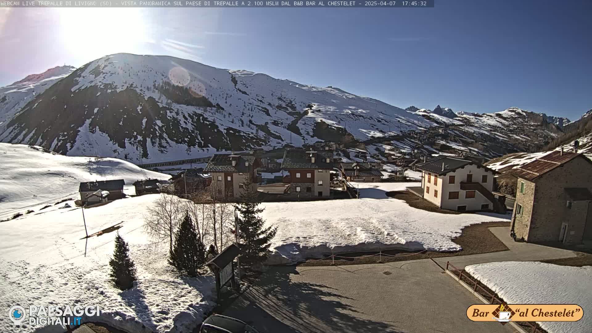A sunny, snow-covered mountain village nestled in a valley, with several buildings and patches of bare ground visible.