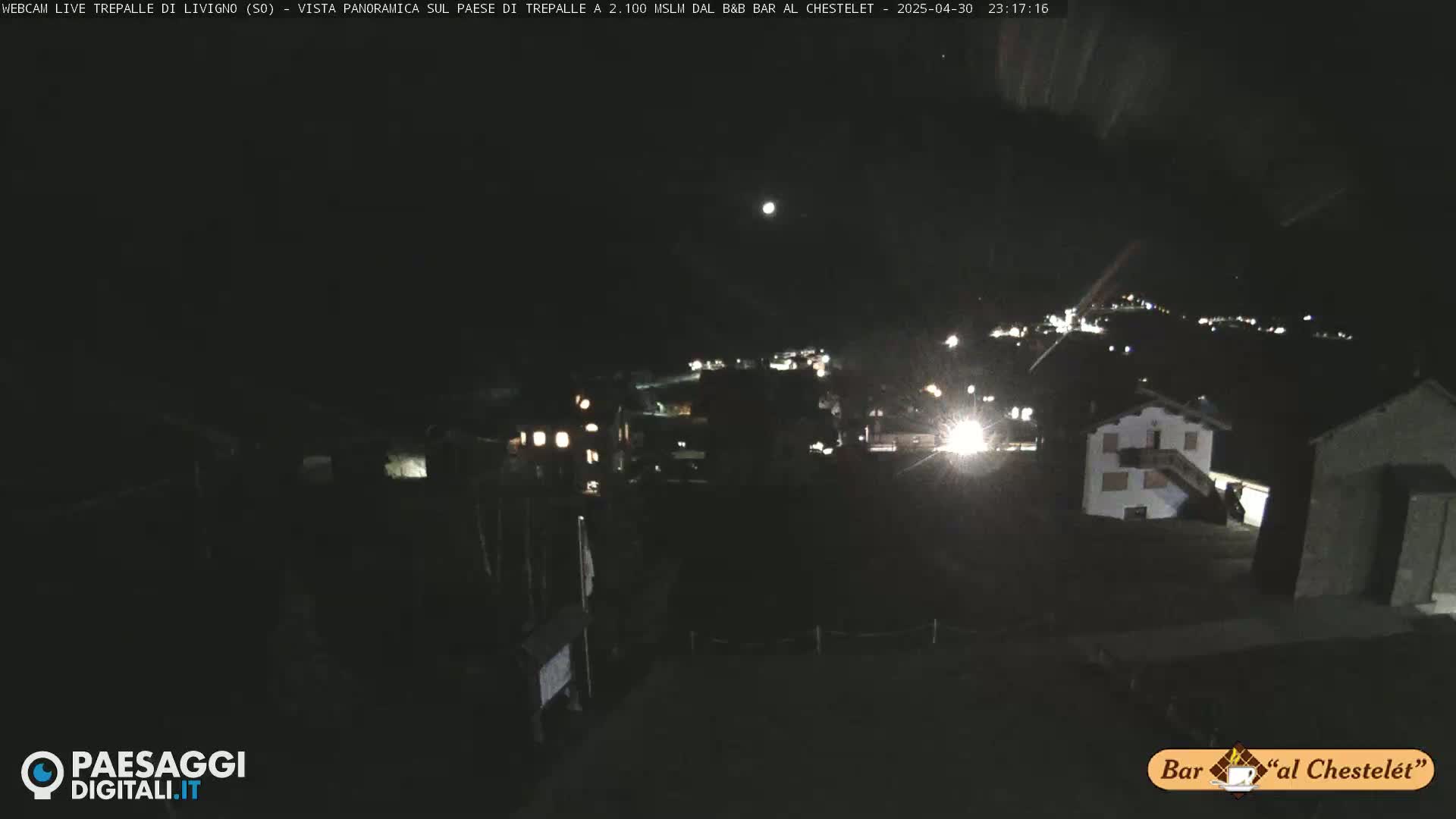 A nighttime, long shot of a village illuminated by scattered lights under a dark sky with a visible moon.