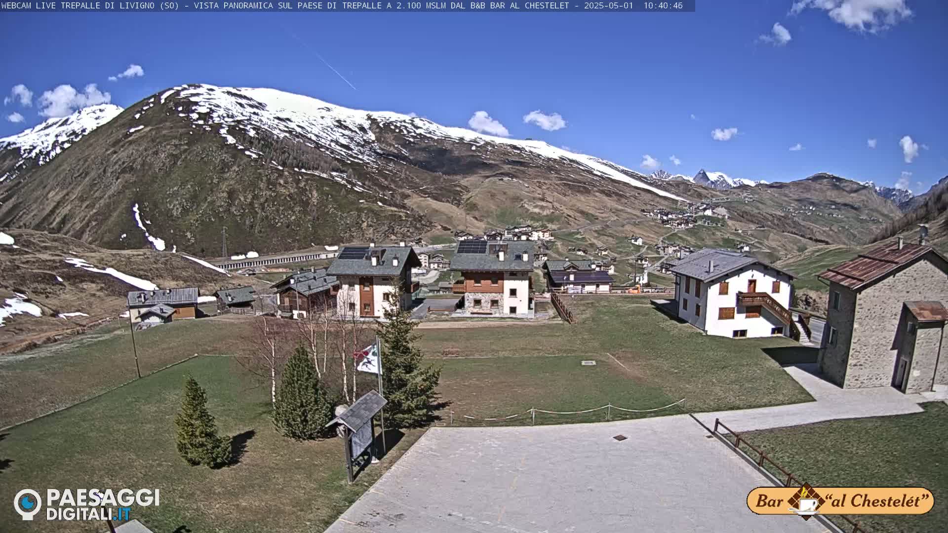 A village nestled in a mountain valley with snow-capped peaks in the background under a mostly sunny sky.