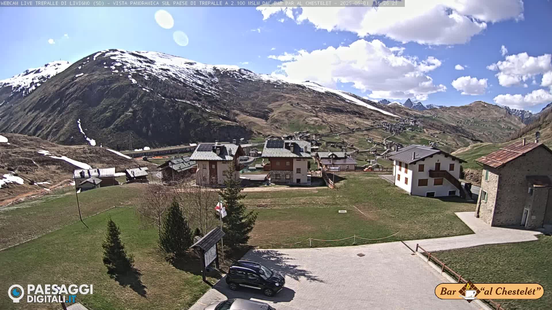 A mountain village with several buildings, a grassy area, and a parking lot sits below a snow-capped mountain under a partly cloudy sky.
