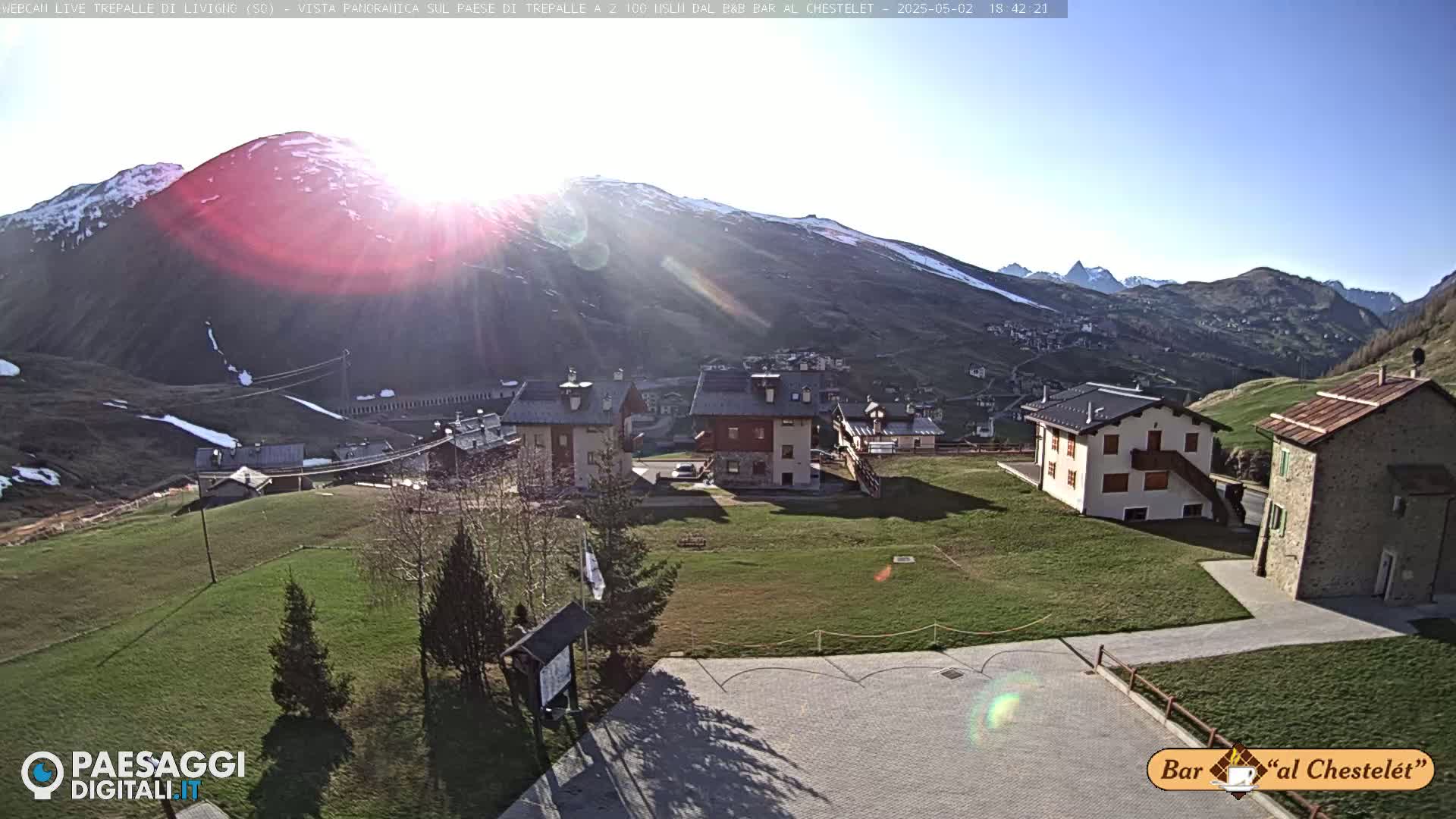 A sunny view of a small village nestled in a mountain valley, with patches of snow visible on the surrounding slopes.